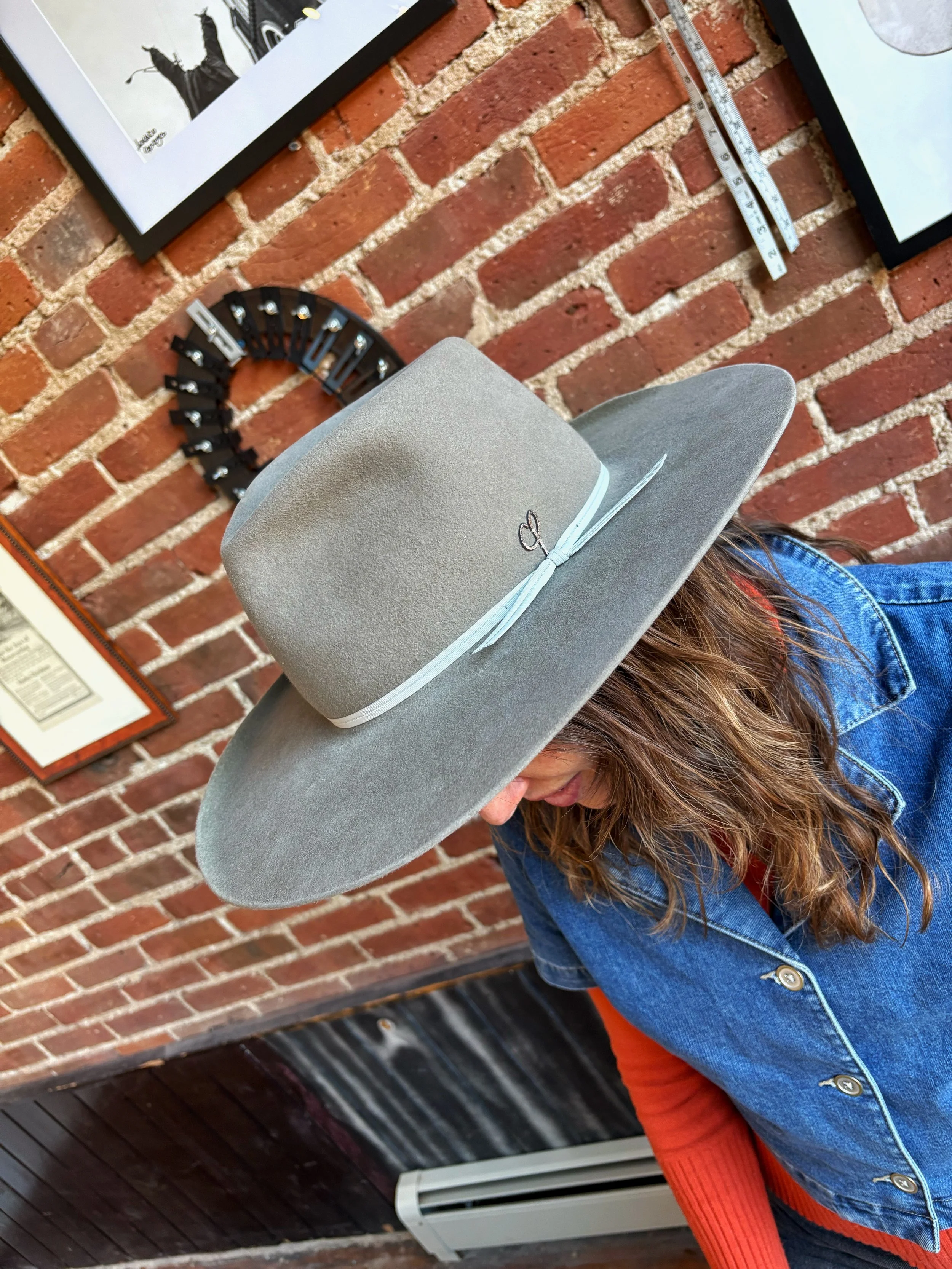 A woman wearing a grey hat with a ribbon bow and a denim jacket, standing in front of a brick wall with framed artwork and a small black decor piece.