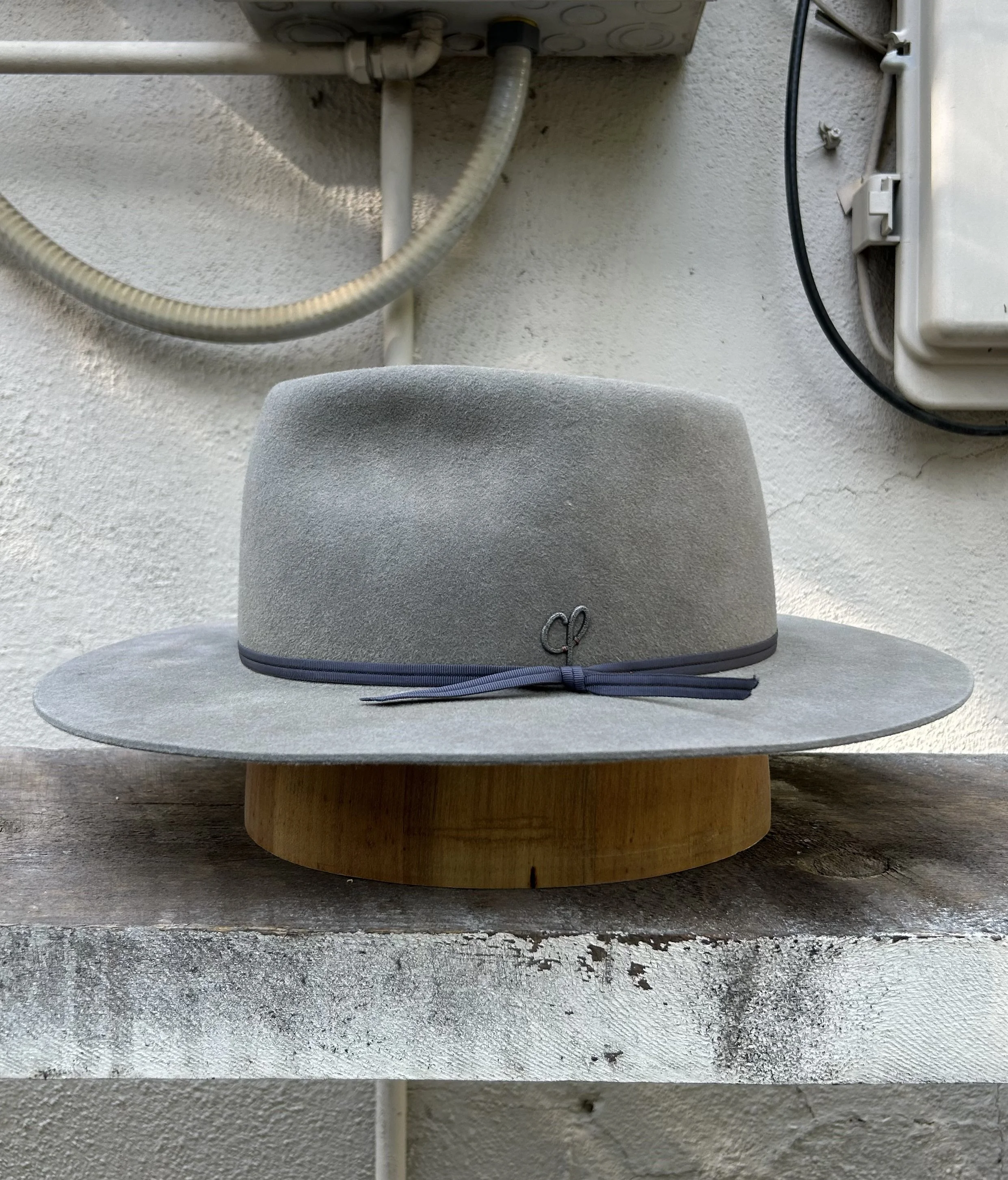 A light gray hat with a black ribbon placed on a wooden stand on a concrete surface, with a textured white wall and some electrical equipment in the background.