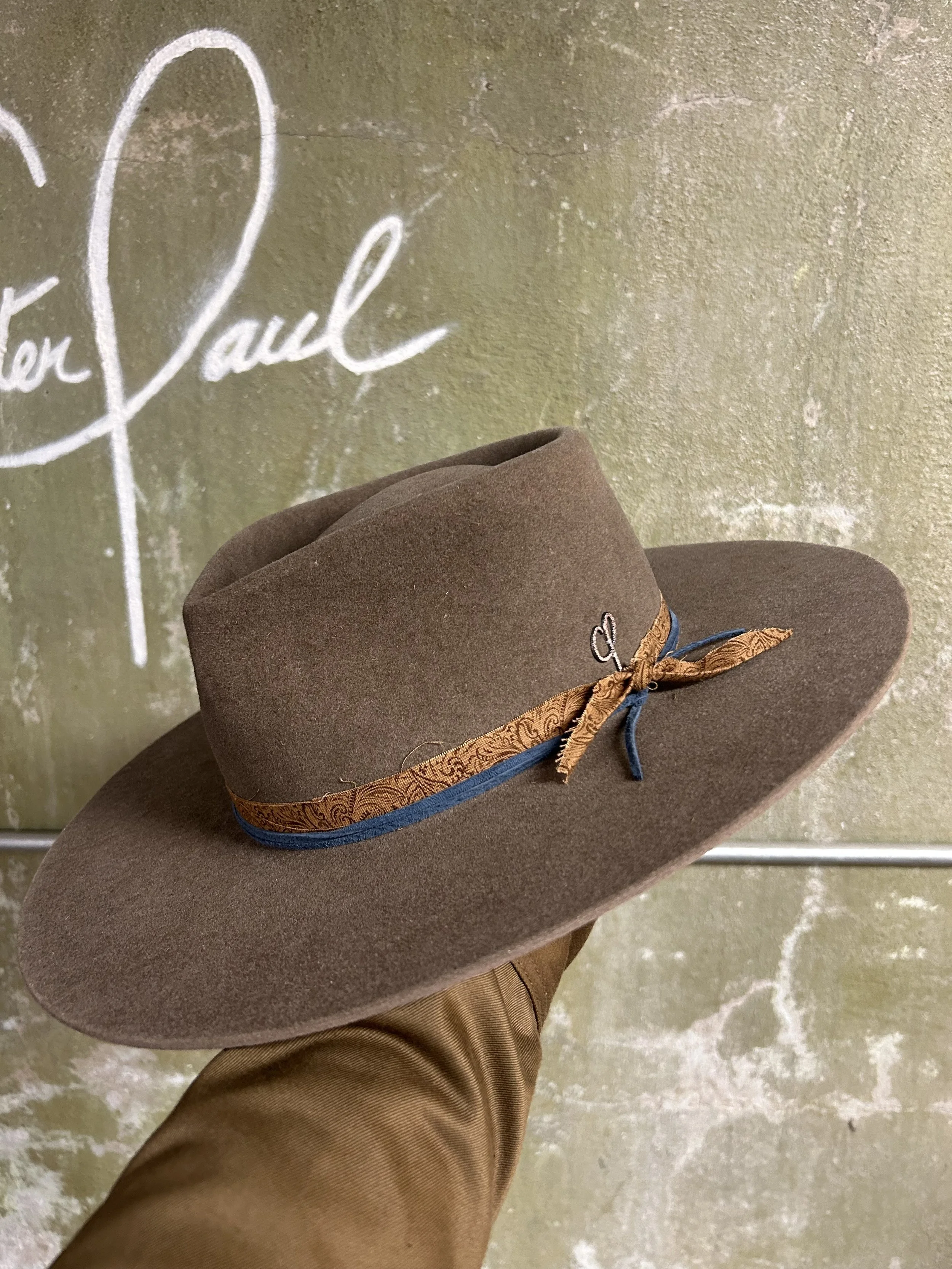 A brown wide-brimmed hat with a decorative band and bow, being held in front of a textured green wall with white script writing.