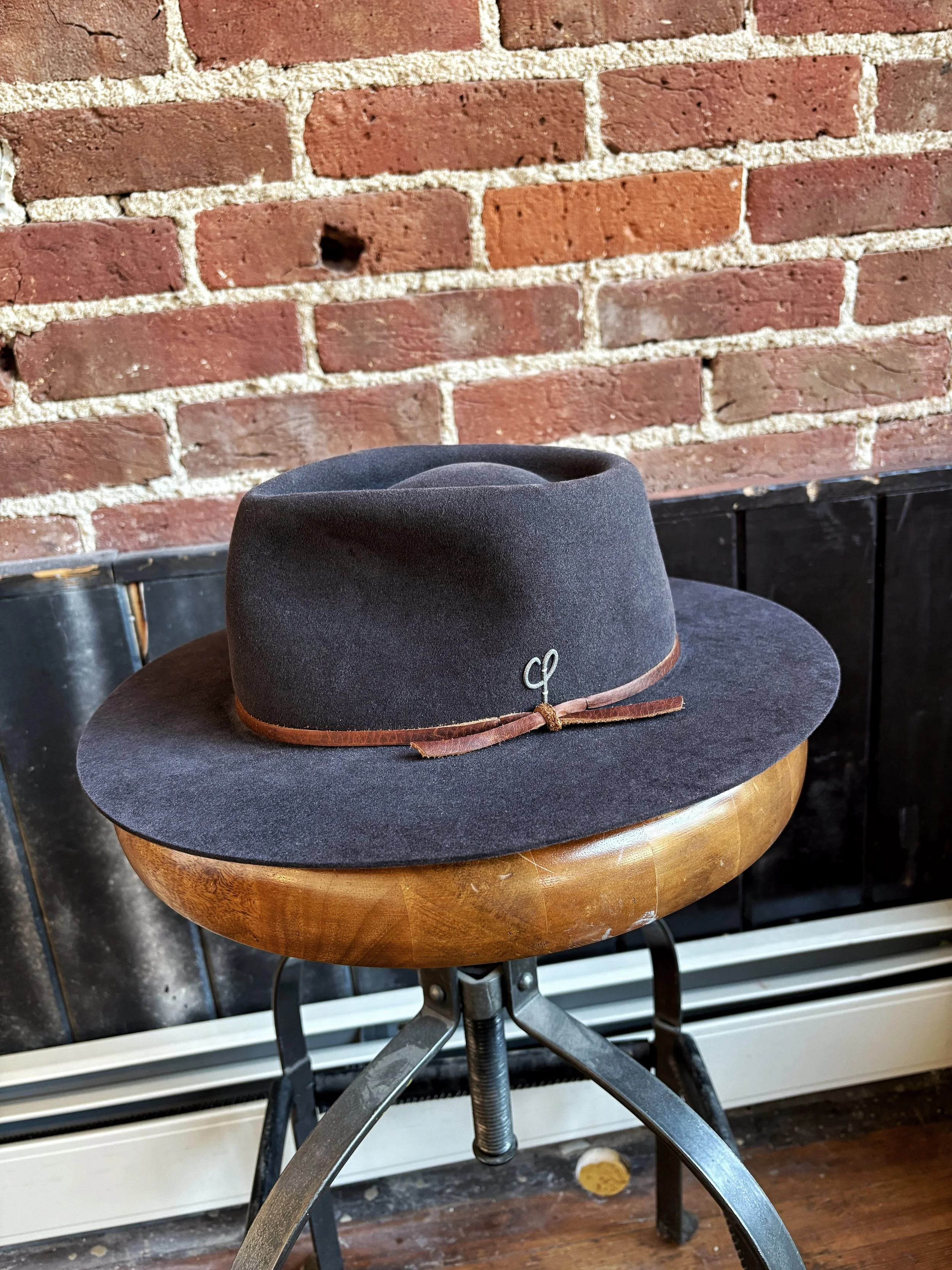 A dark gray fedora hat with a brown leather band and a metal decorative pin, sitting on a wooden stool against a brick wall background.