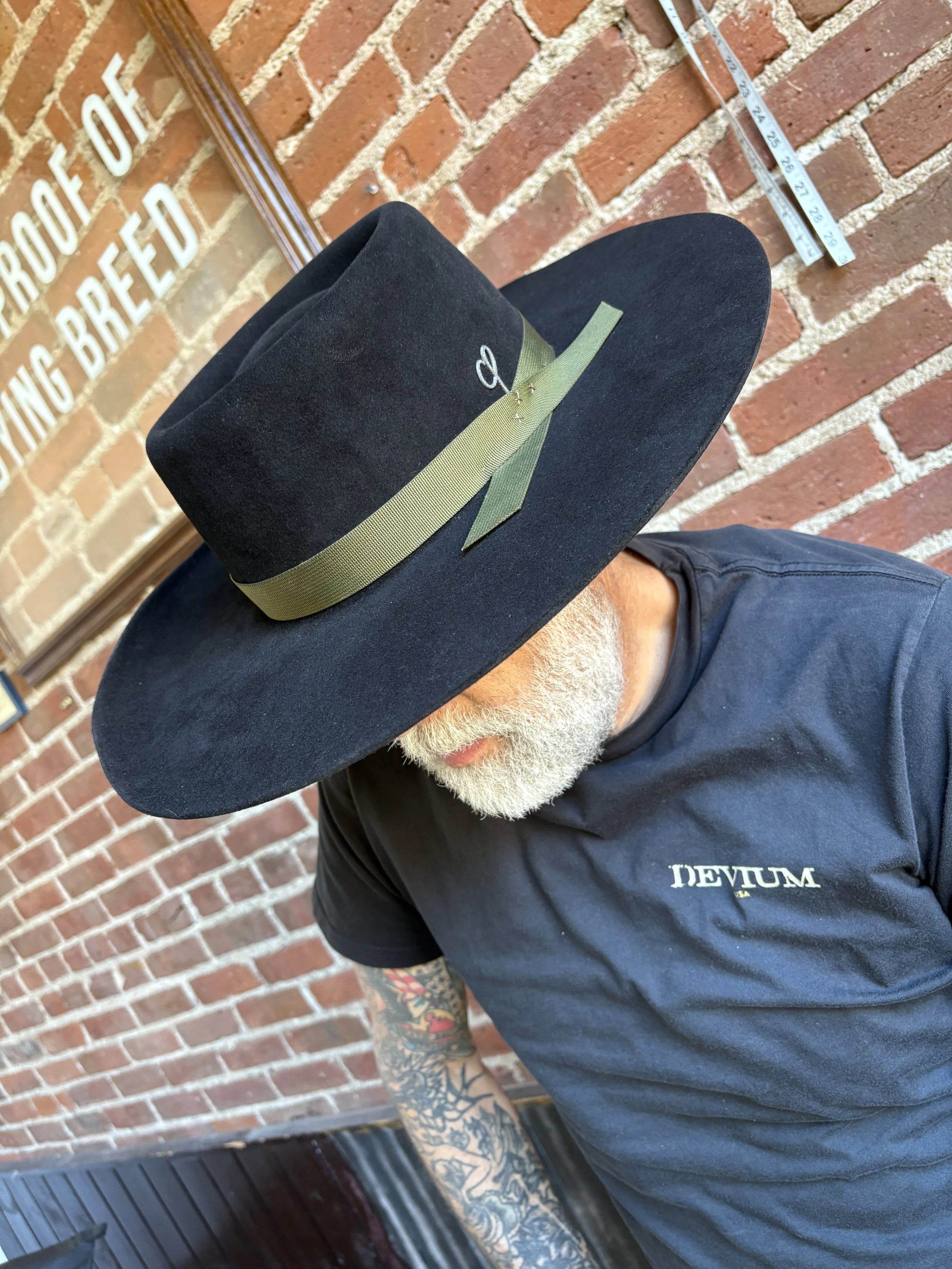 A man with a white beard and tattoos on his arm wearing a black T-shirt with 'DEVIUM' printed on it, and a large black hat with a green ribbon and a pin, sitting against a brick wall.