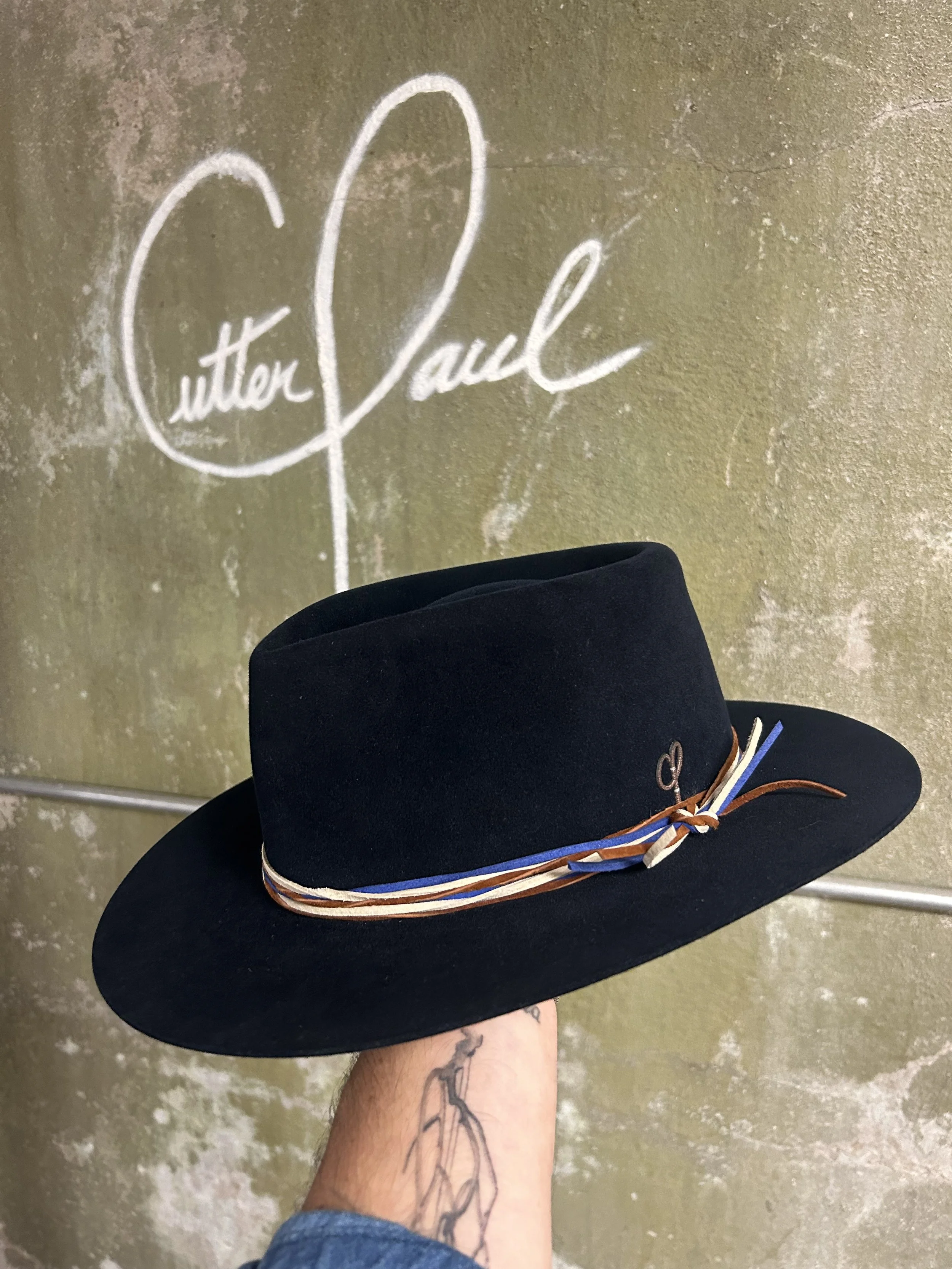A black hat with a colorful band, held by a tattooed hand, in front of a wall with the 'Christian Siriano' signature in white.