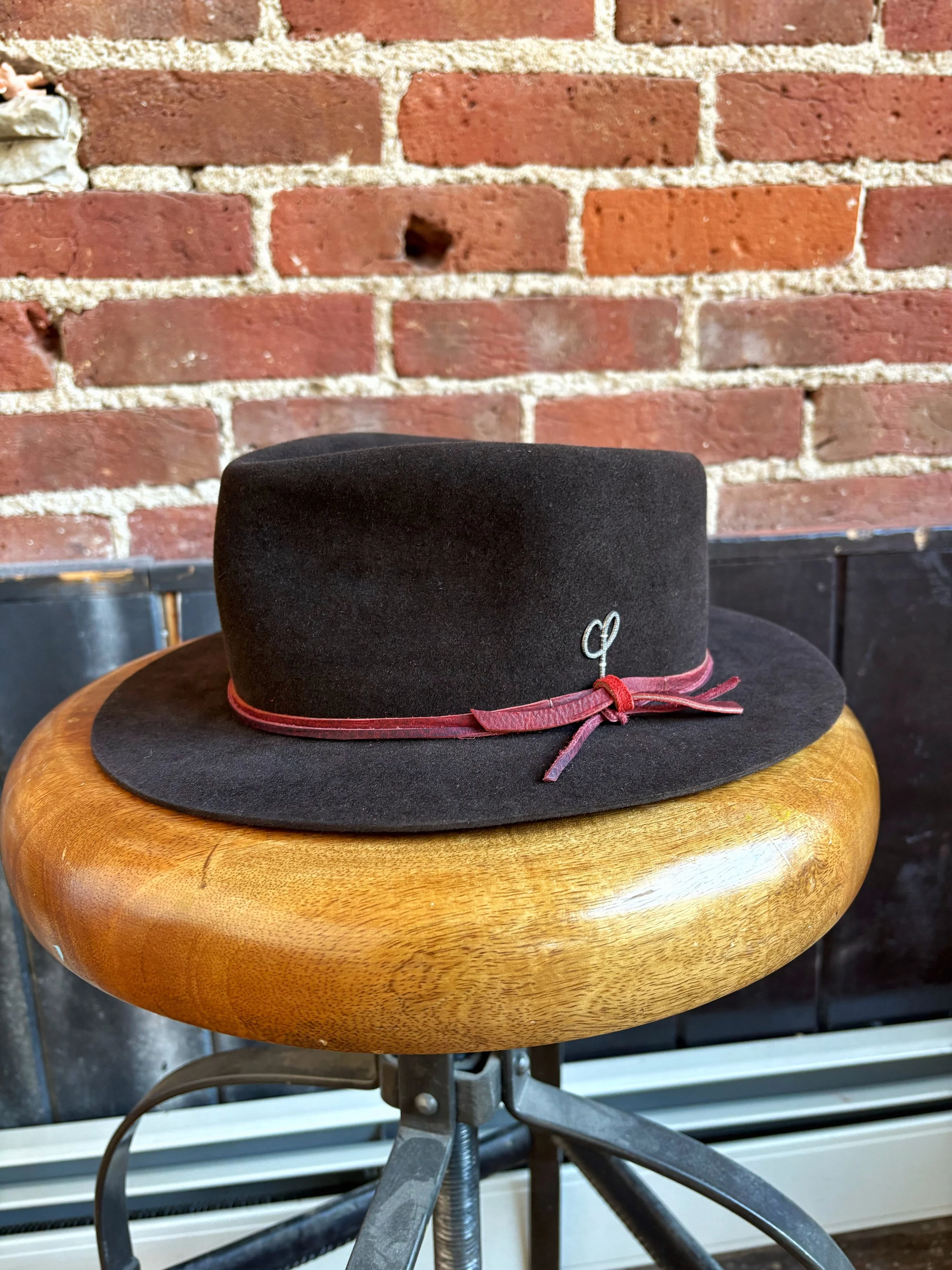 A black felt fedora hat with a red band and a small silver pin, placed on a round wooden stool with a curved seat, against a brick wall background.