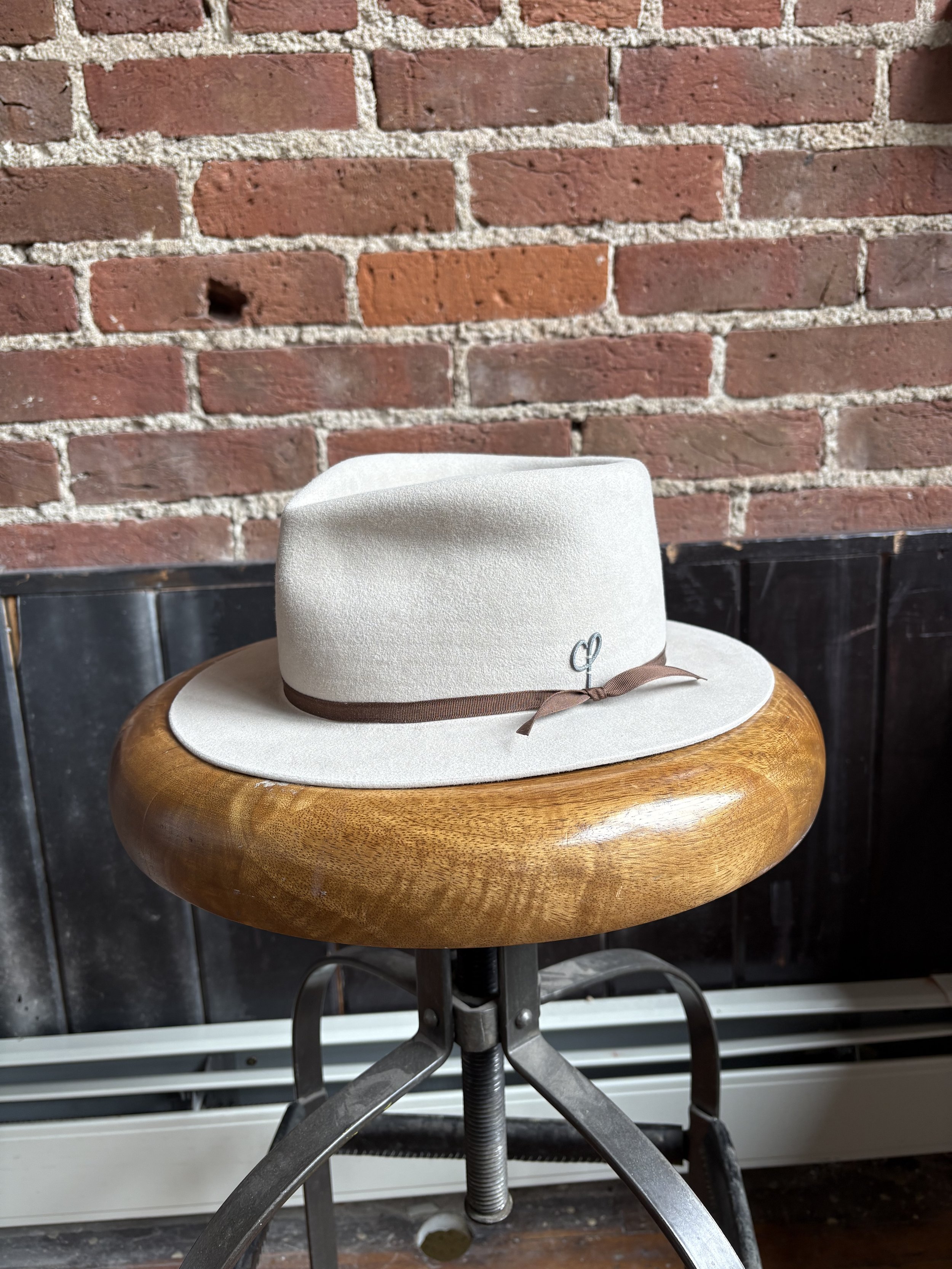 A white felt fedora hat with a brown ribbon and a small silver charm on a wooden stool against a brick wall background.