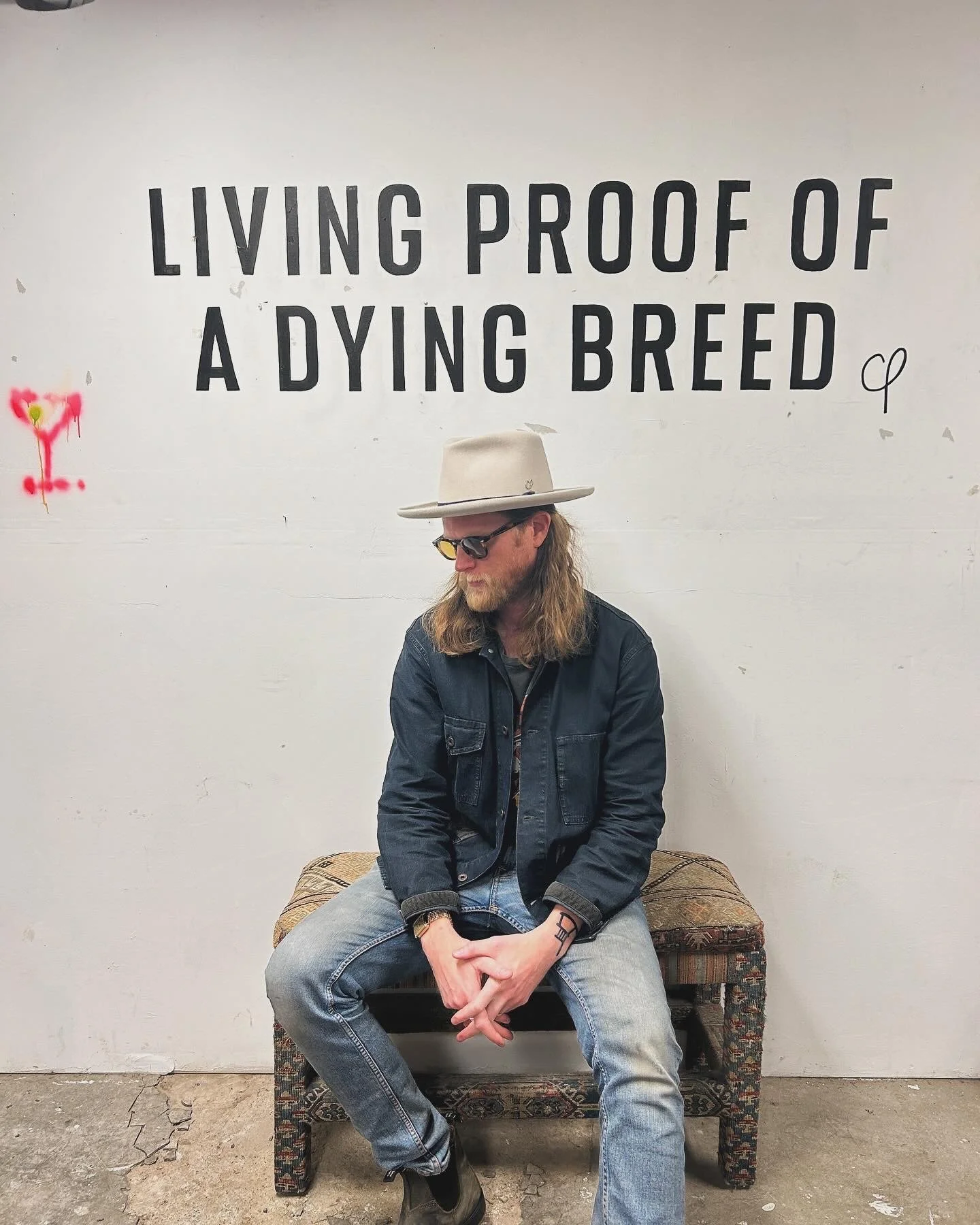 A man with long hair, beard, sunglasses, a beige hat, a black denim jacket, and jeans sits on a patterned bench in front of a white wall with black text that says, 'Living Proof of a Dying Breed.'