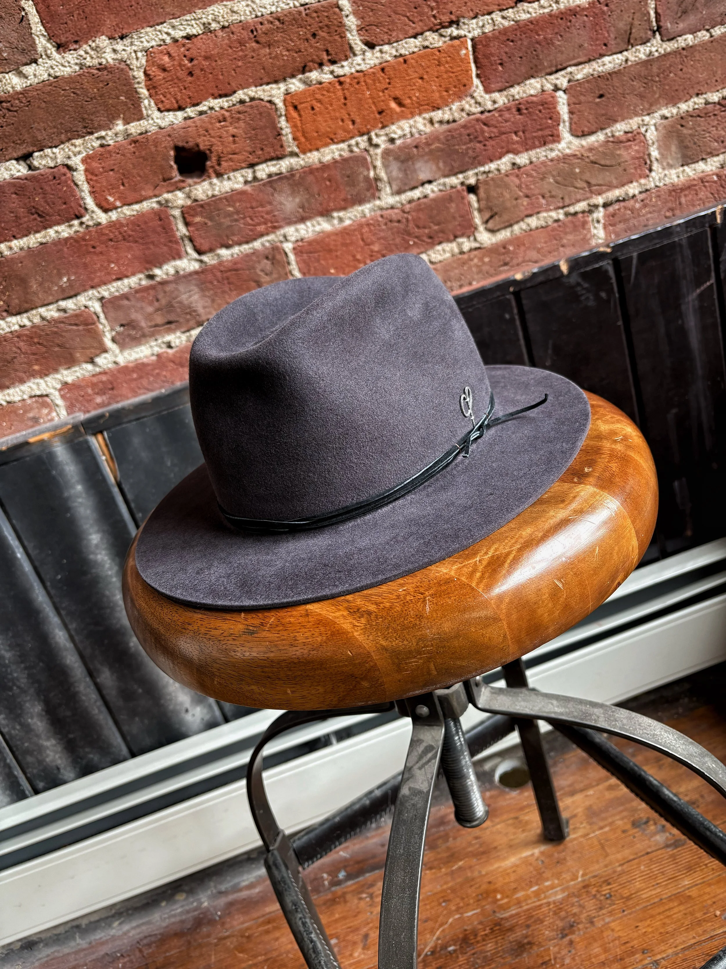 A grey fedora hat with a black band resting on a round wooden stool with metal legs, against a brick wall background.