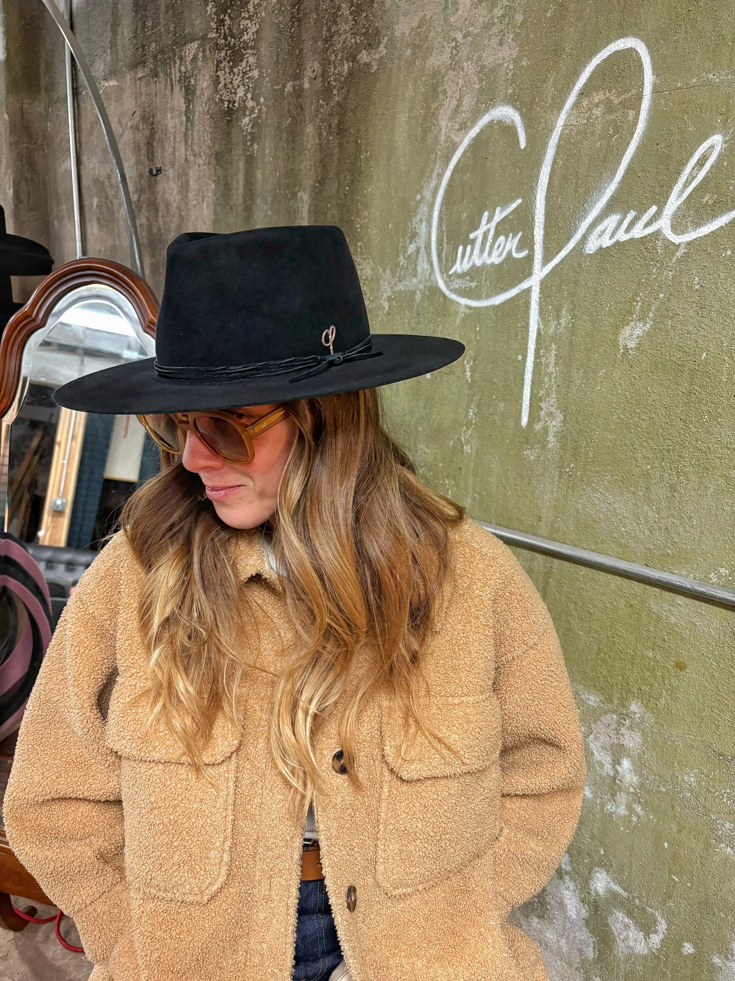 A woman with wavy hair wearing a black wide-brimmed hat, sunglasses, a tan fleece jacket, and blue jeans, standing in front of a green wall with white cursive writing.