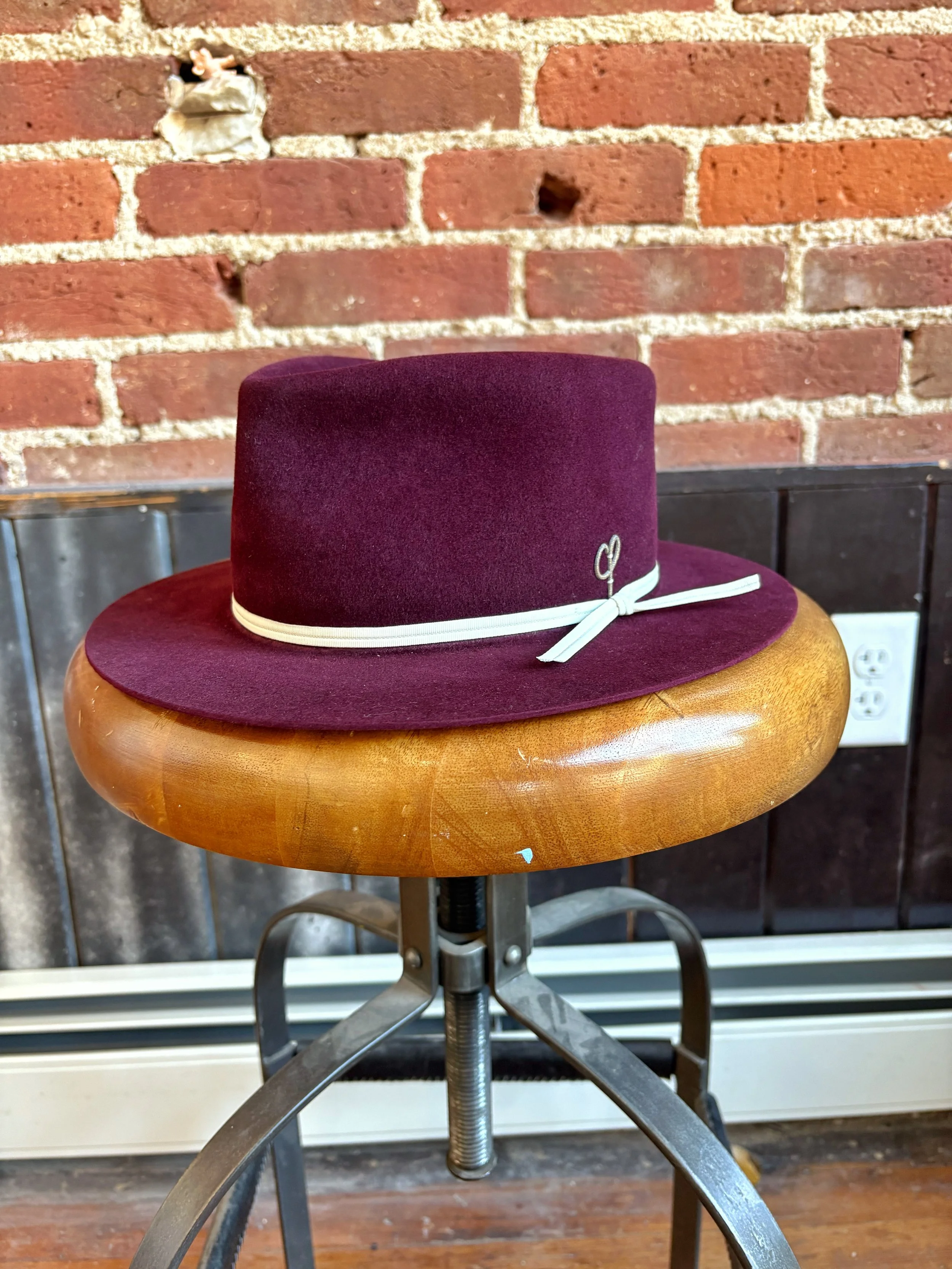 A burgundy felt hat with a white ribbon, placed on a wooden stool in front of a brick wall.