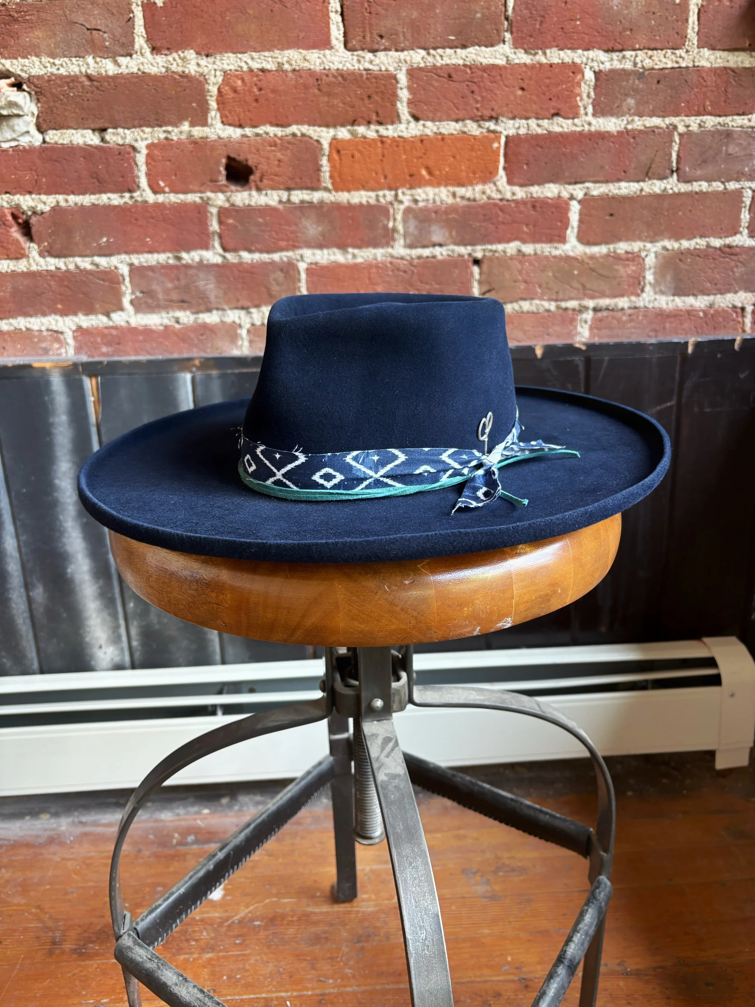 A navy blue fedora hat with a band featuring geometric patterns, placed on a round wooden-topped adjustable stool with a metal base, against a brick wall background.