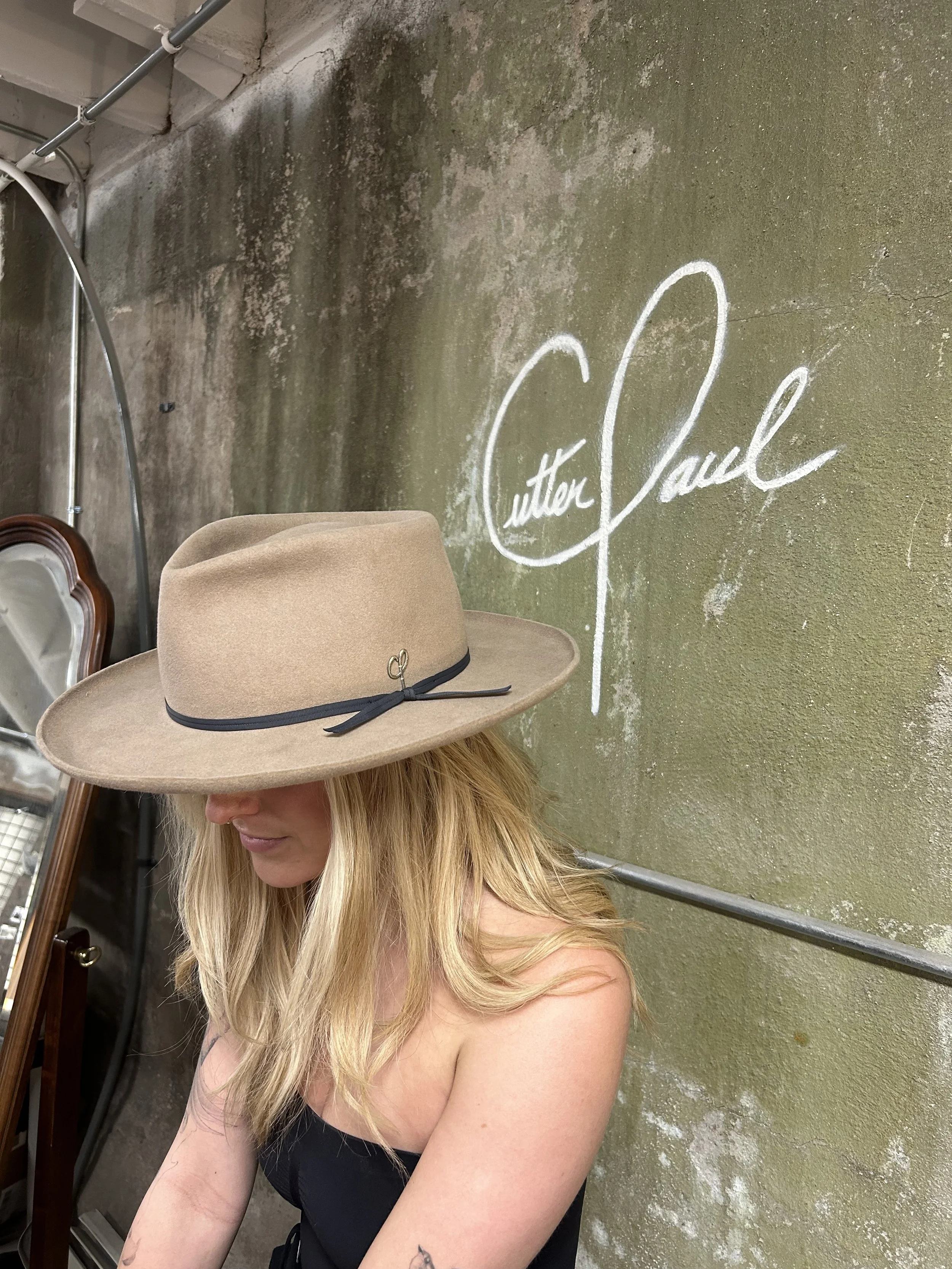 A woman with blonde hair wearing a beige wide-brimmed hat with a black ribbon, standing in front of a textured, mossy green wall with white chalk writing that reads 'Cutter Paul'.