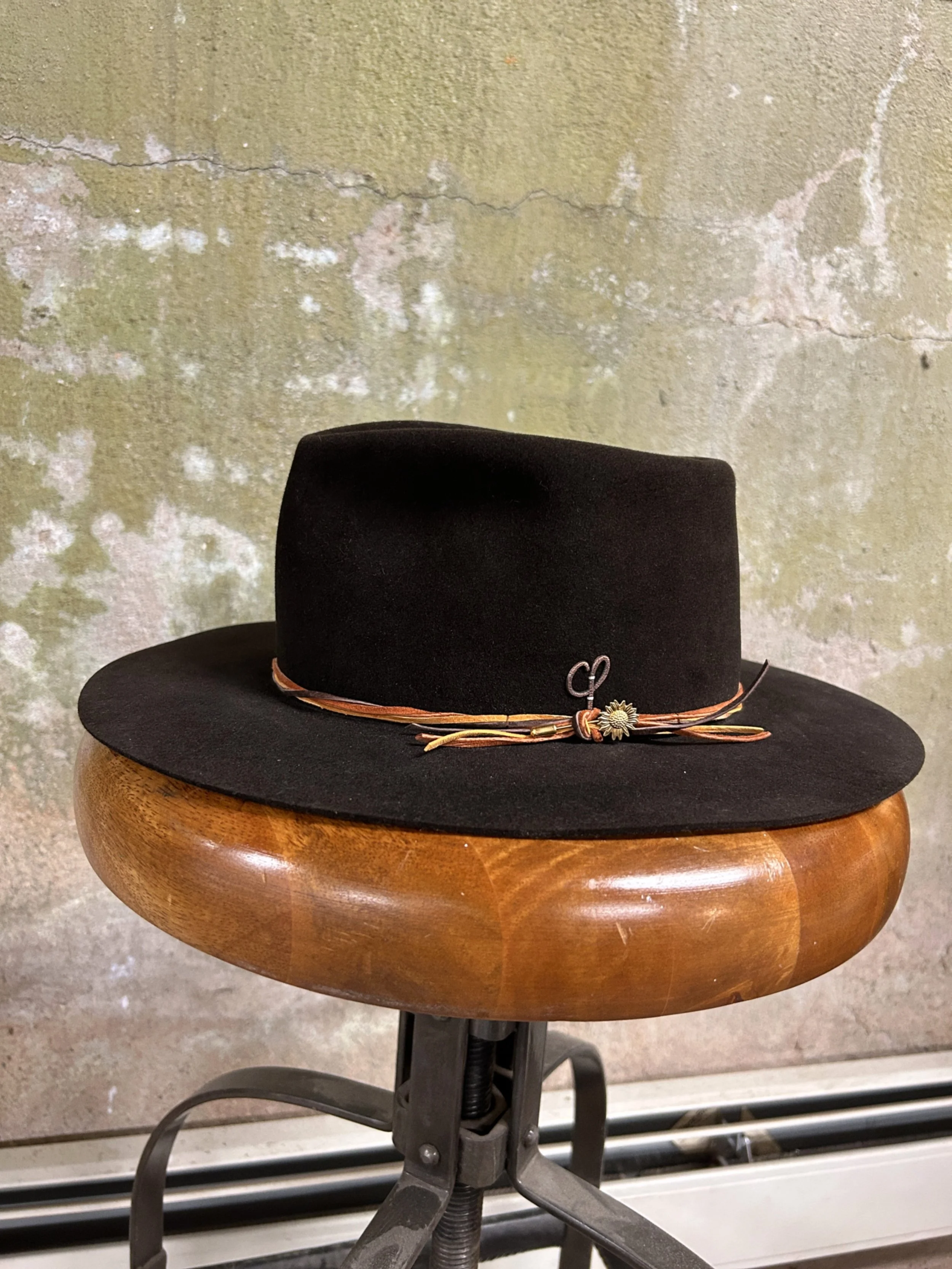 Black wide-brimmed hat with a leather band and decorative pin, placed on a wooden saddle, against a textured wall.