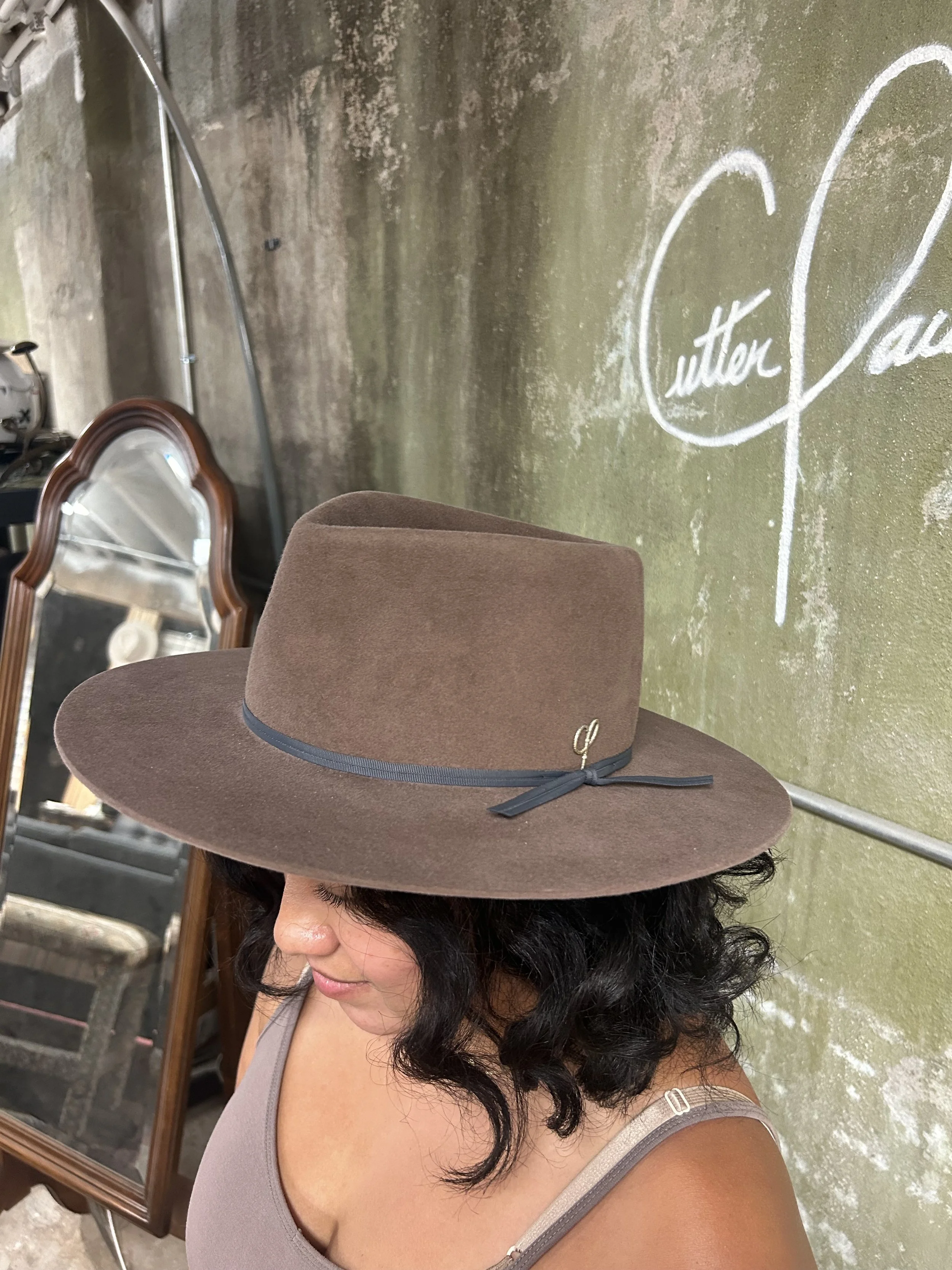 A woman wearing a large brown hat with a black band and a small gold logo, standing indoors near a mirror and a green wall with white chalk writing.