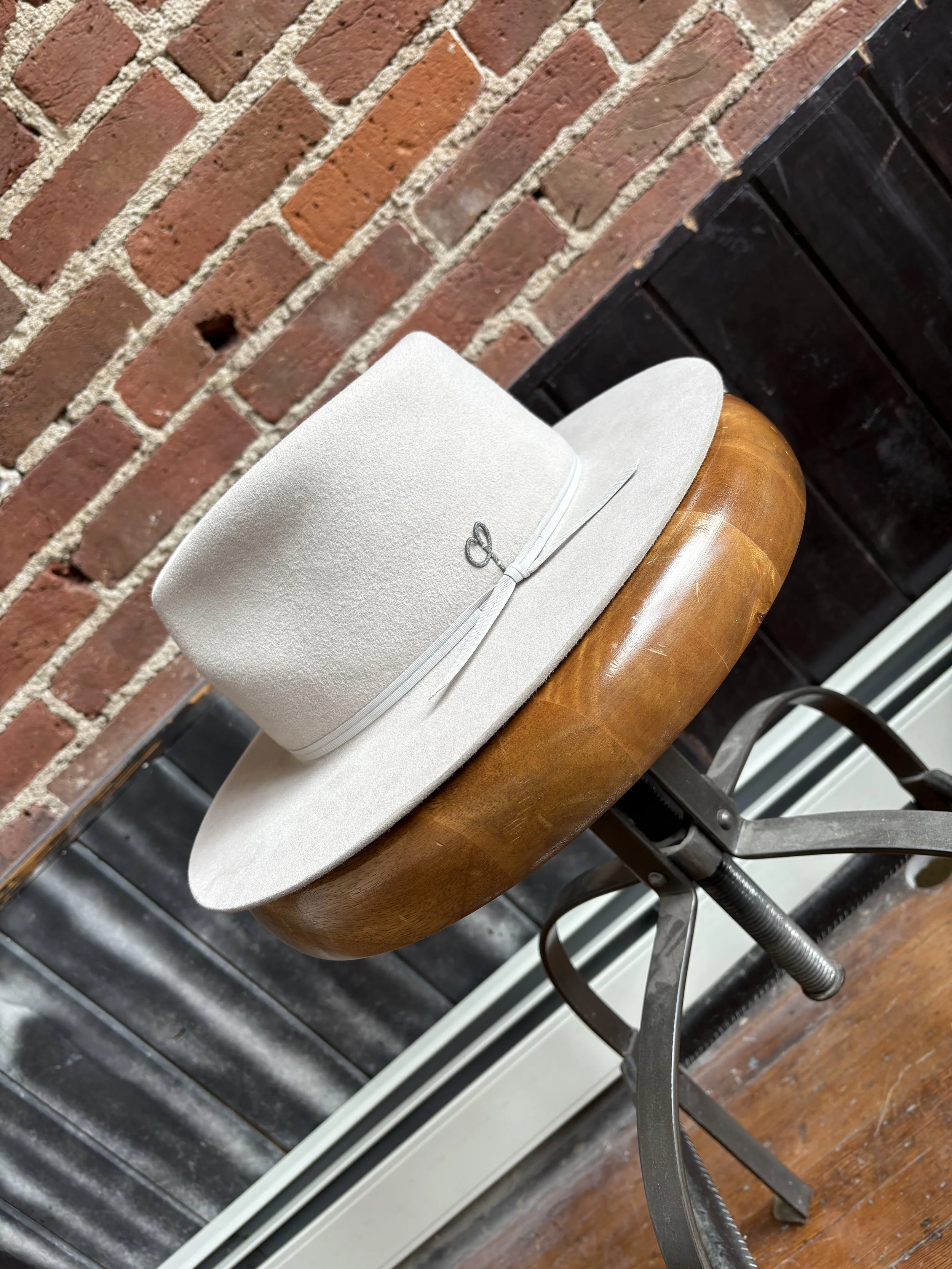A white hat with a ribbon and bow, resting on a beige felt hat box, placed on a wooden candelabra-style stand against a brick wall background.