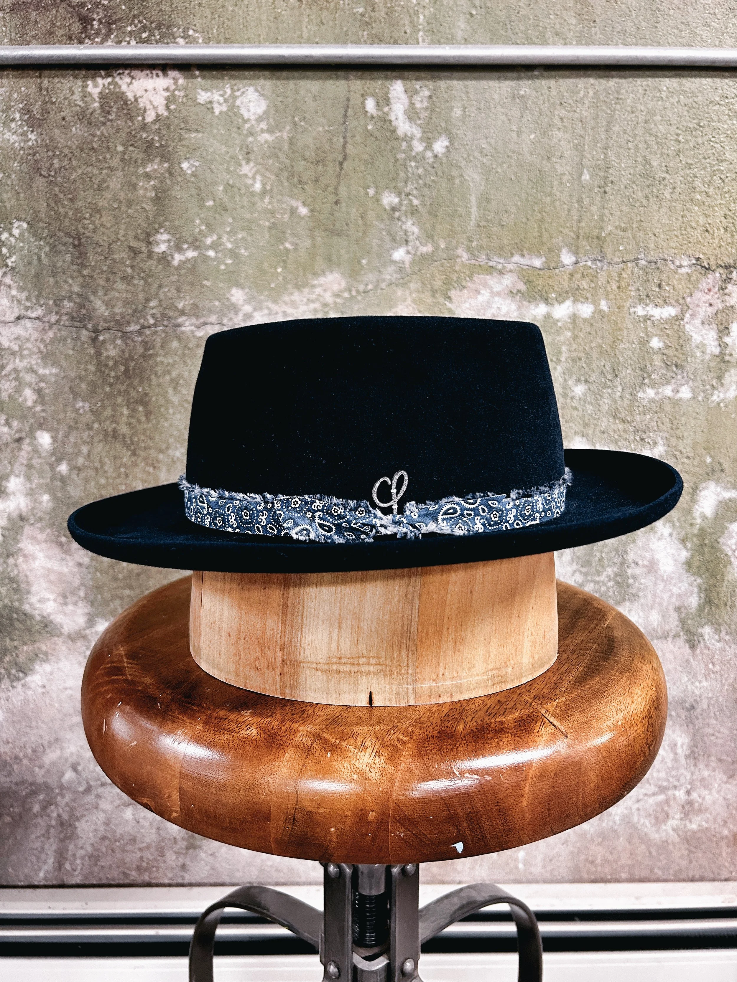 Black wide-brimmed hat with a blue patterned band on an orange wooden stool against a textured wall background.