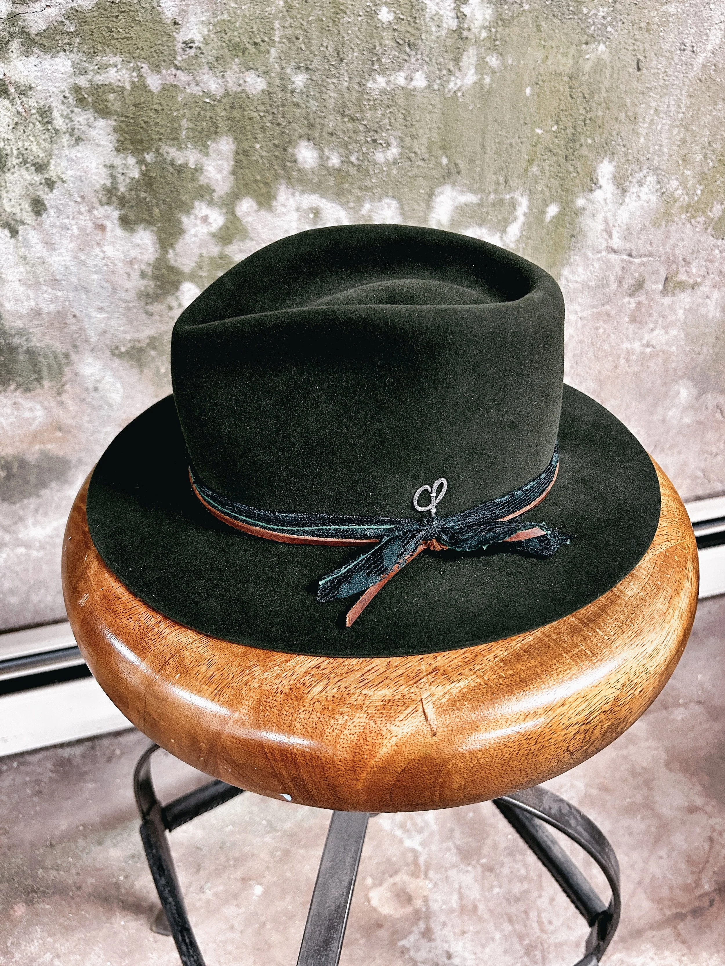 Black felt fedora hat with a ribbon and small silver clasp, placed on a wooden stool against a textured wall background.