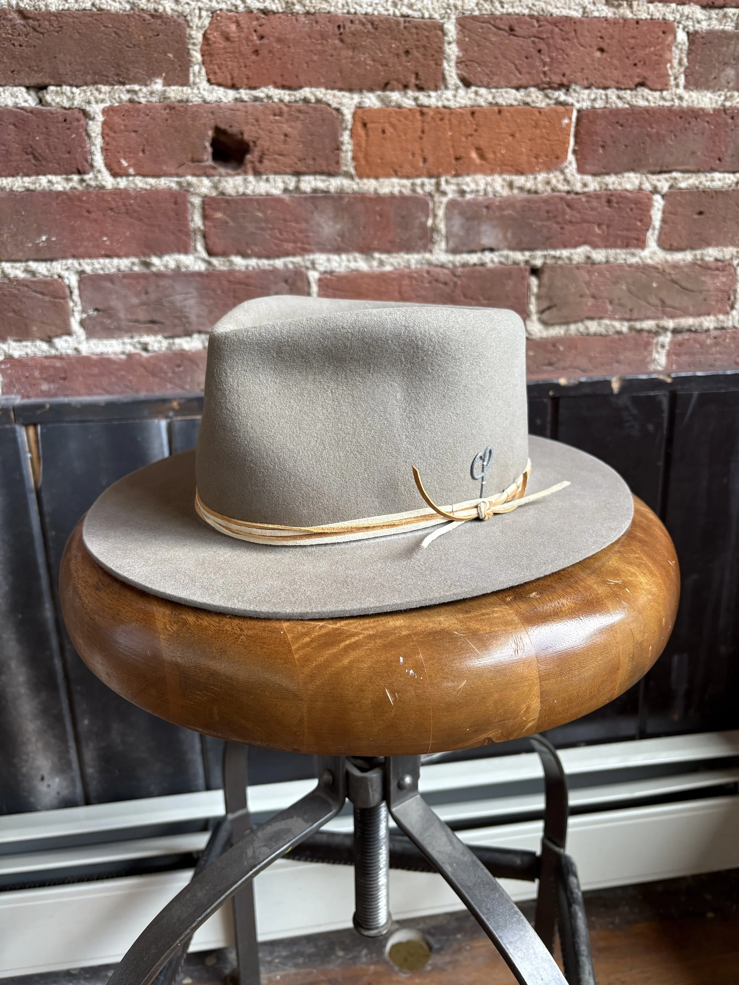 A light gray wide-brimmed hat with a beige band and a small metal detail, placed on a round wooden stool against a brick wall background.