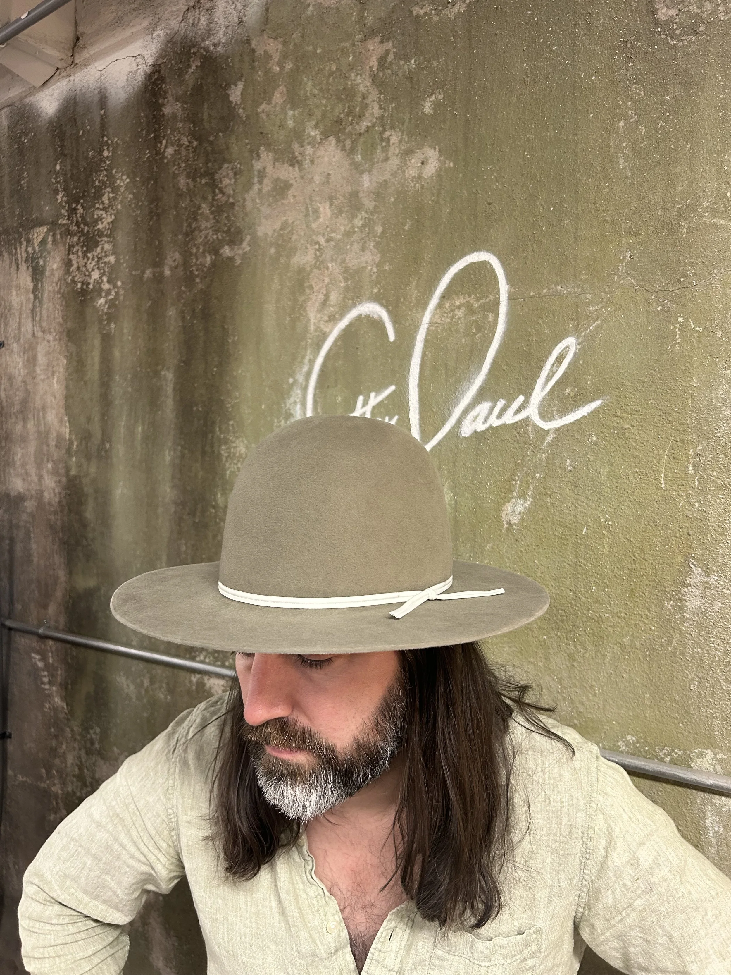A man with long hair and a beard wearing a beige shirt and a wide-brimmed beige hat with a white band, standing in front of a wall with a painted signature that says 'C. Pauch'.