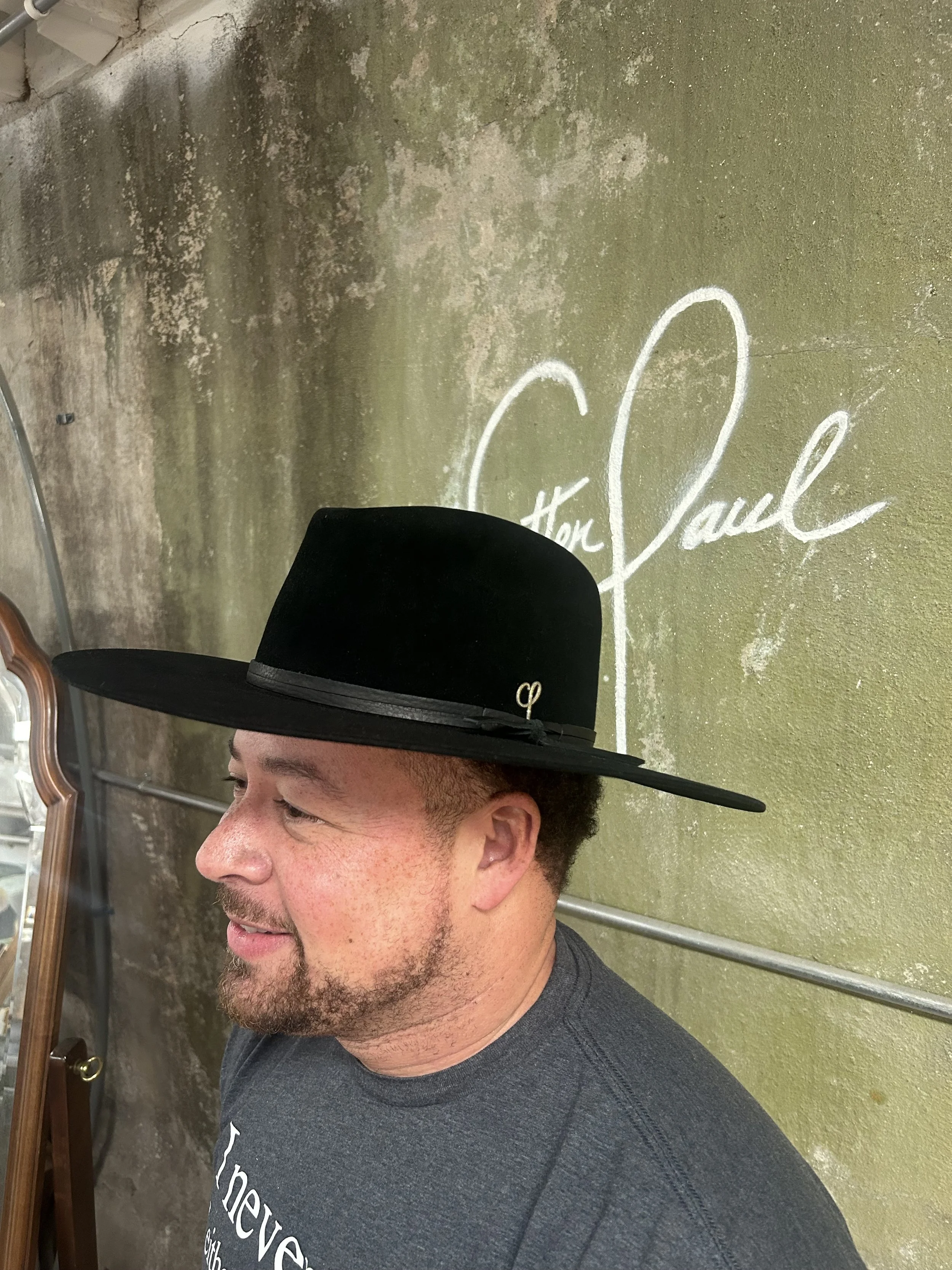 A man wearing a large, black wide-brim fedora hat with a small metal logo, standing in front of a mossy, textured wall with the name 'St. Paul' written on it in cursive.