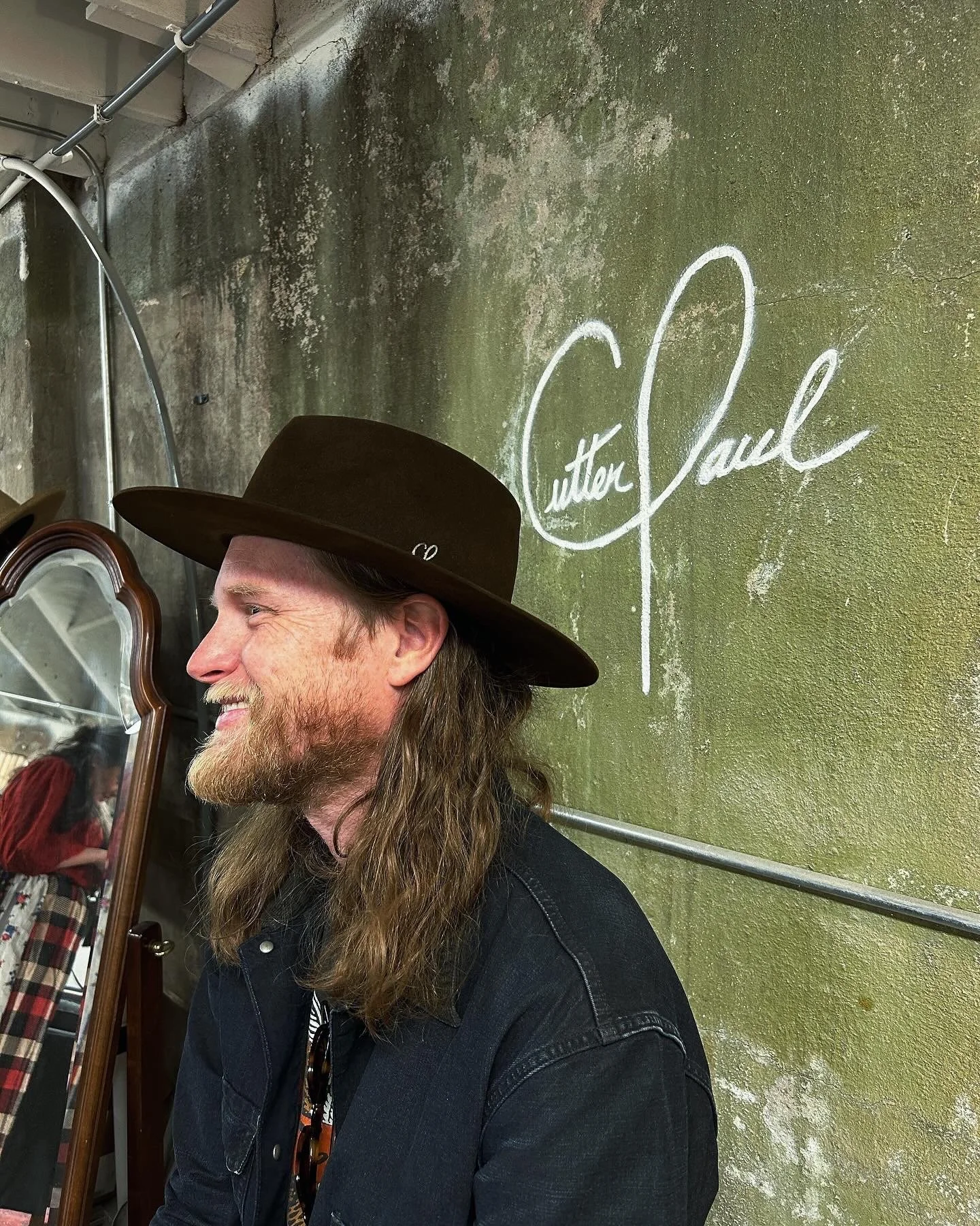 A man with long hair, beard, and a wide-brimmed hat smiling in front of a green wall with a signature that reads 'Cutter J. Paul'.
