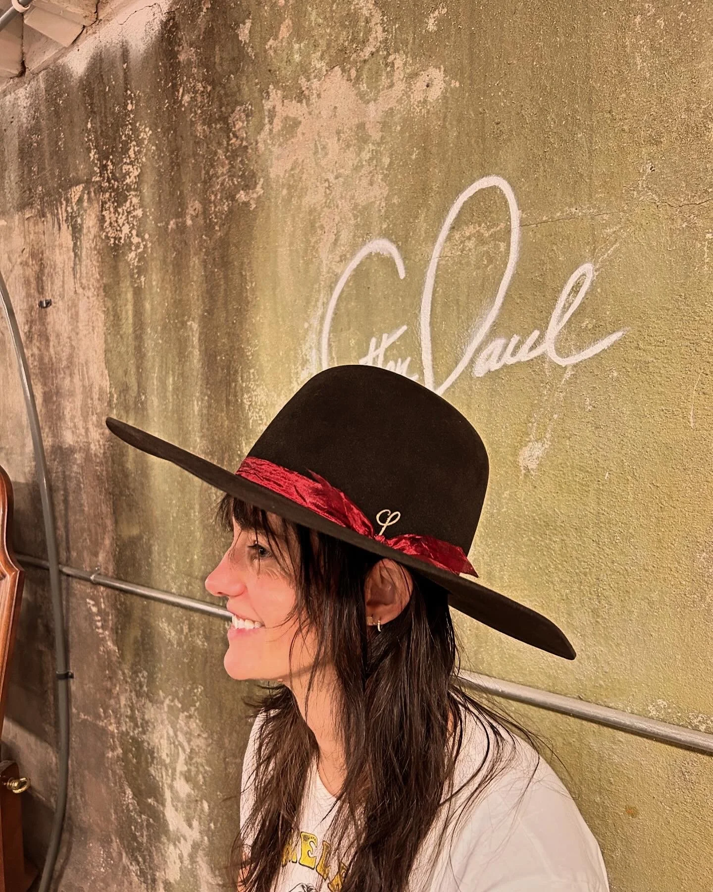 Woman with long brown hair smiling, wearing a large black wide-brimmed hat with a red ribbon, standing against a textured greenish wall with white cursive writing.
