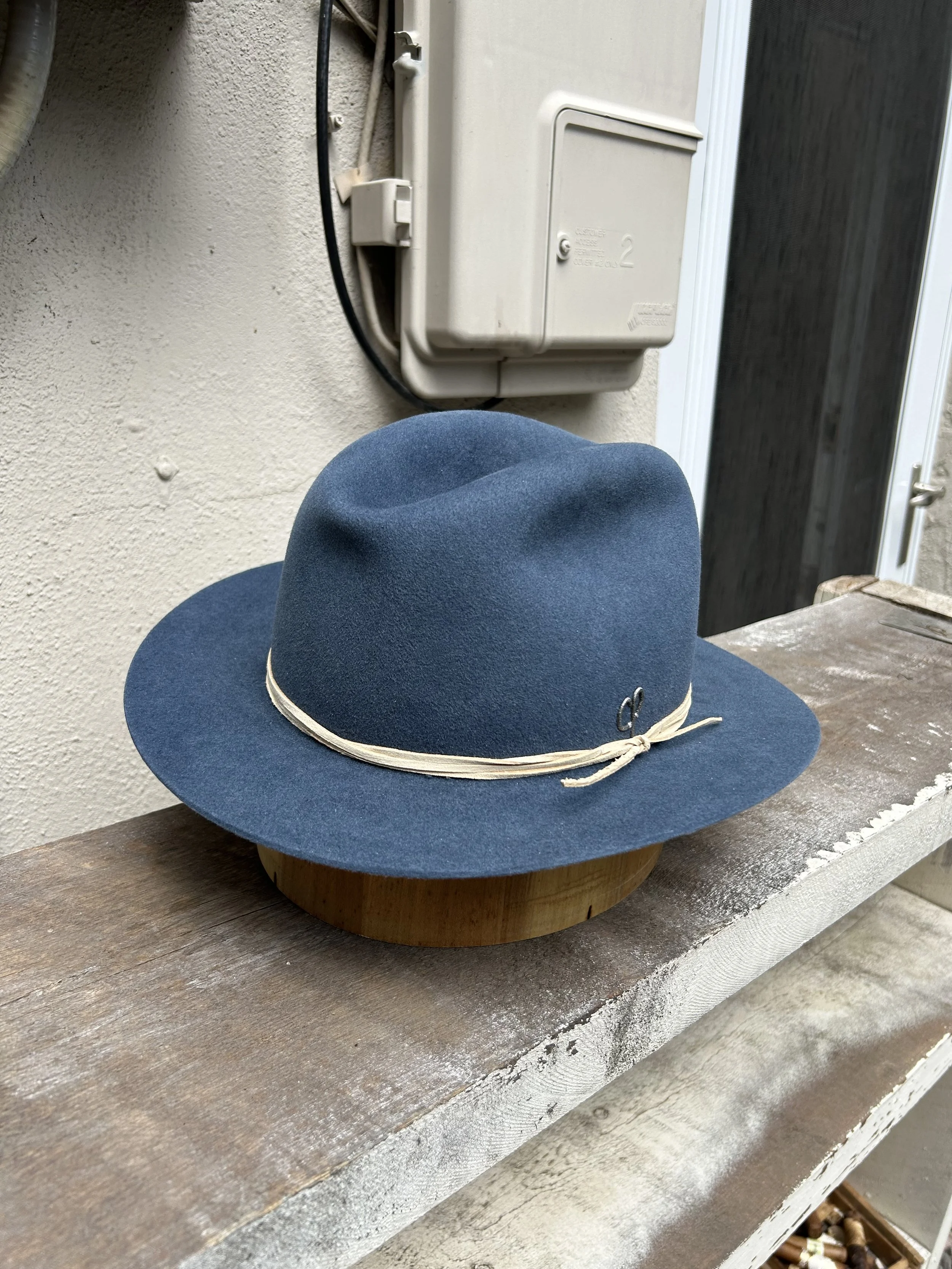 A blue wide-brimmed fedora hat with a beaded band, resting on a wooden stand on a weathered shelf outdoors.
