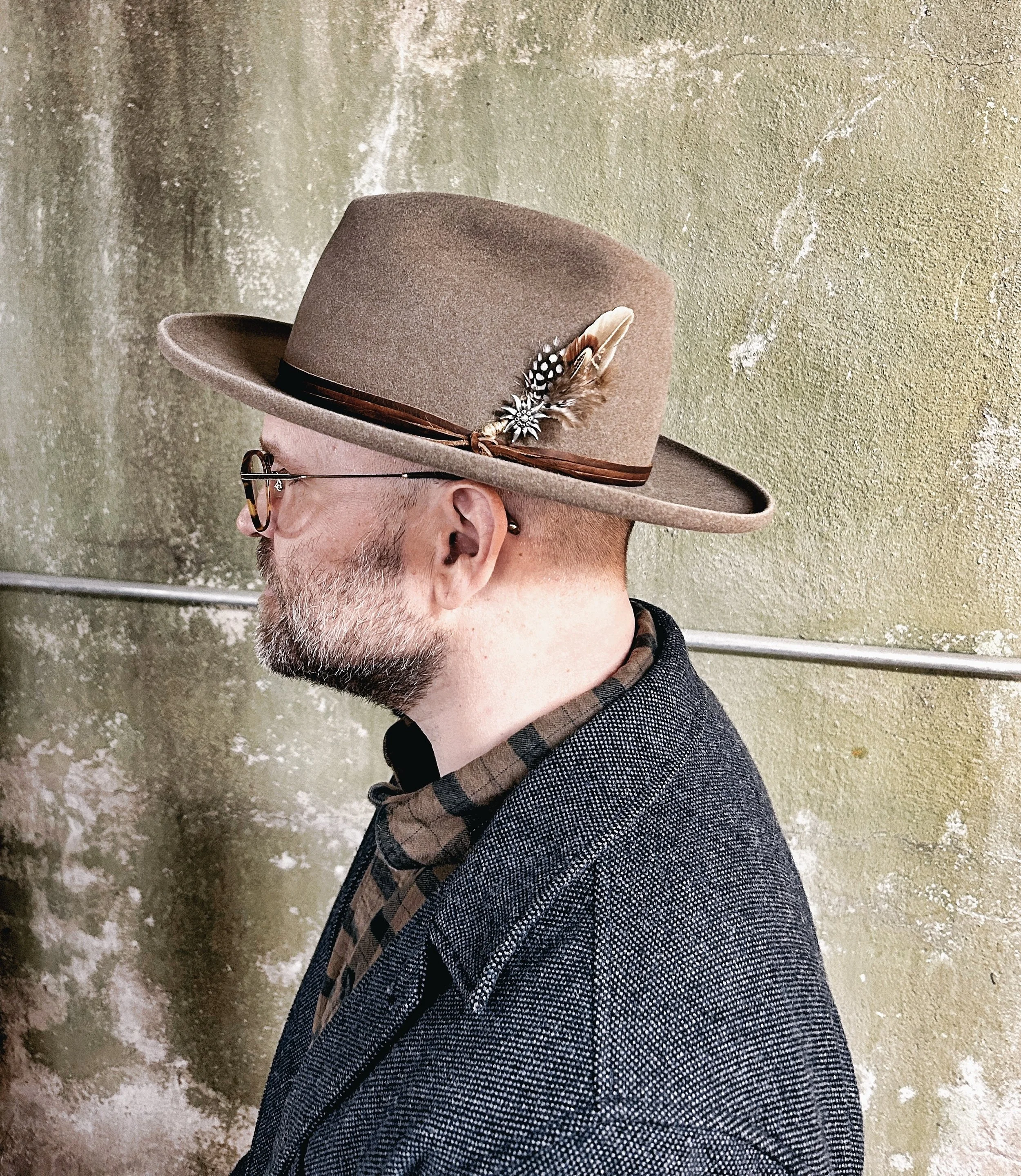 A side profile of a man wearing a brown fedora hat with feathers and beads, glasses, a gray blazer over a plaid shirt, standing against a textured green wall.