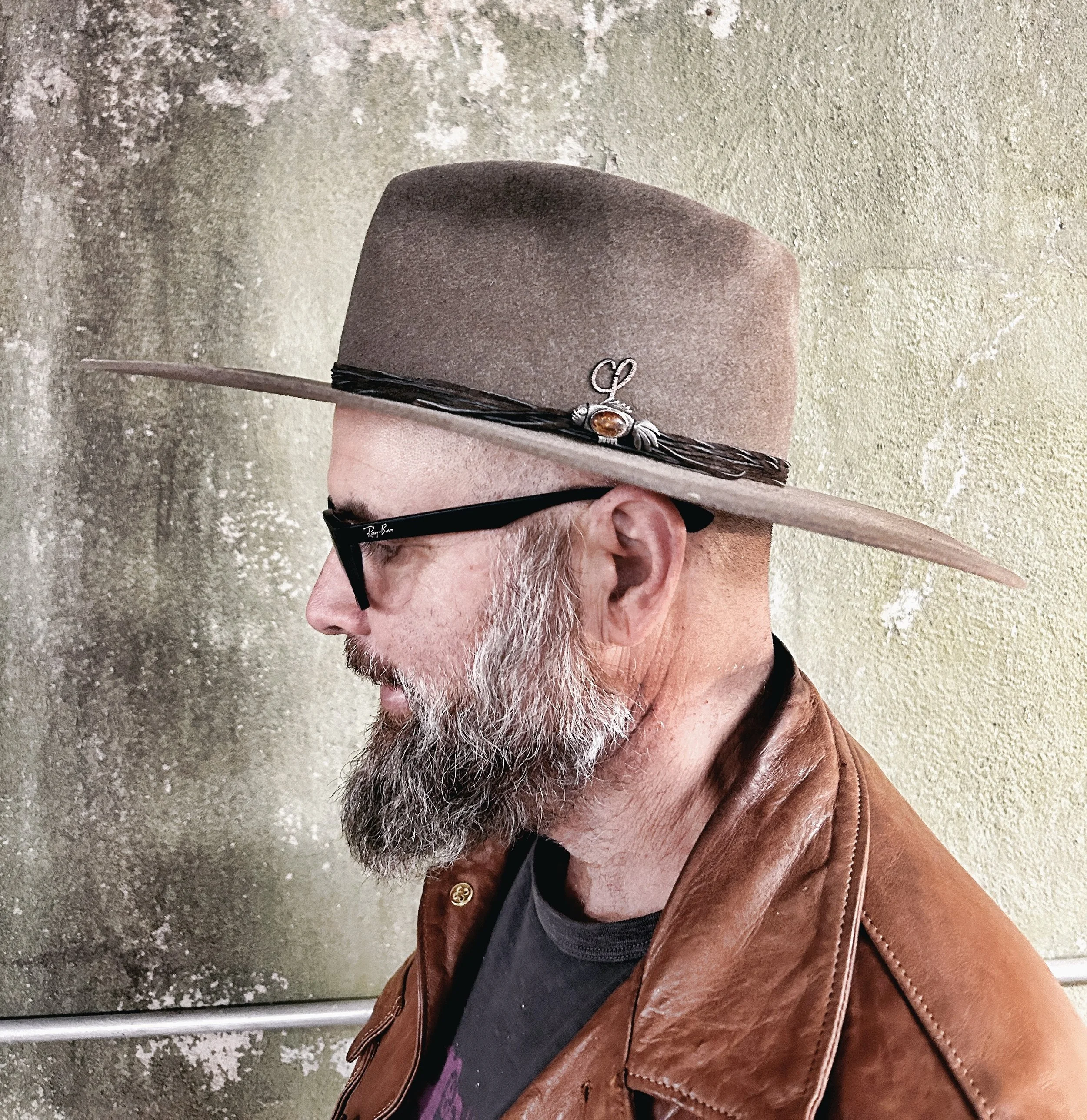 Profile of a man with a gray beard wearing a brown leather jacket, black glasses, and a wide-brimmed hat with decorative pins, standing against a textured wall.