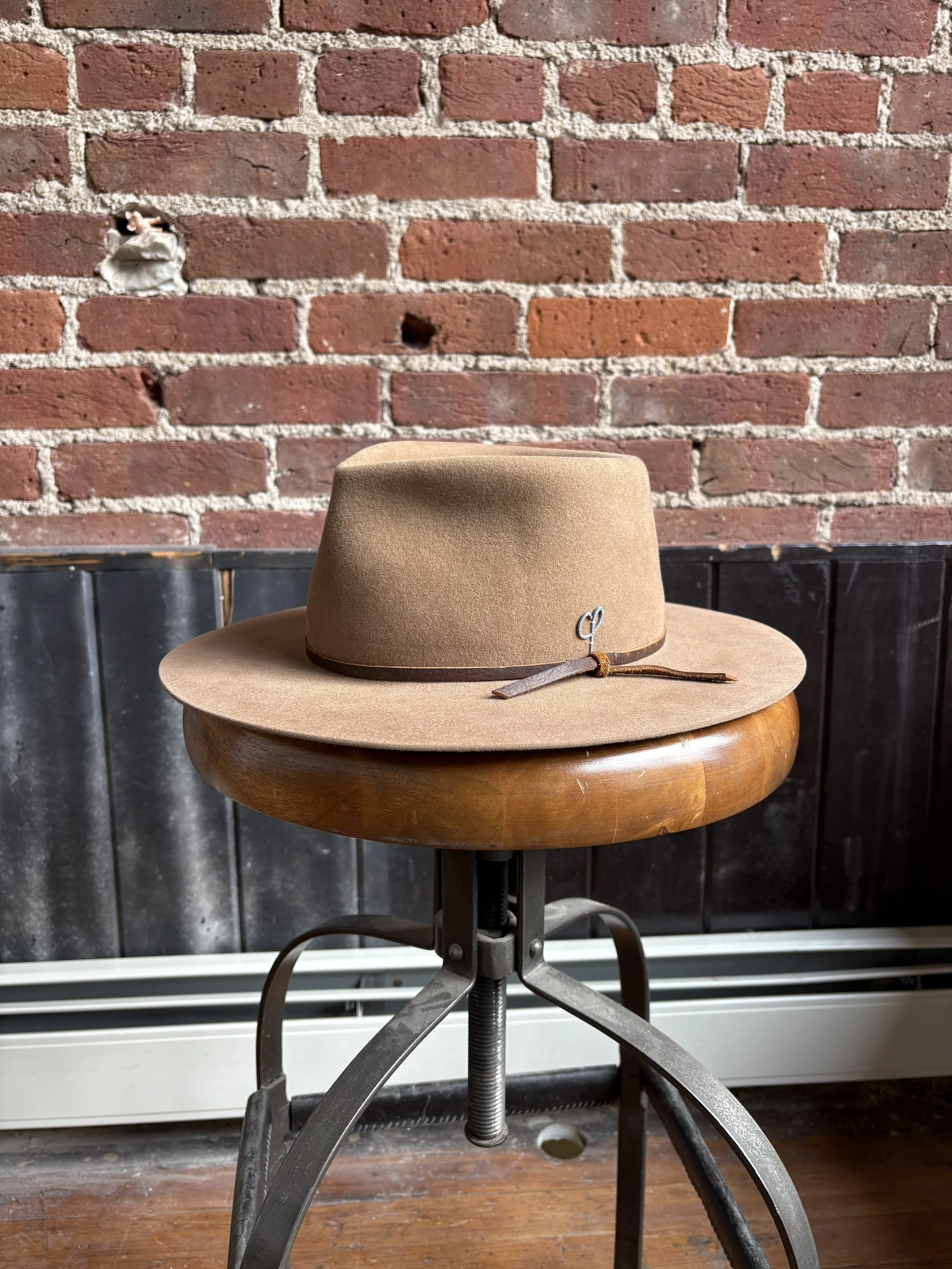 A tan fedora hat with a leather band and a small metal emblem sitting on a wooden and metal adjustable stool in front of a brick wall.