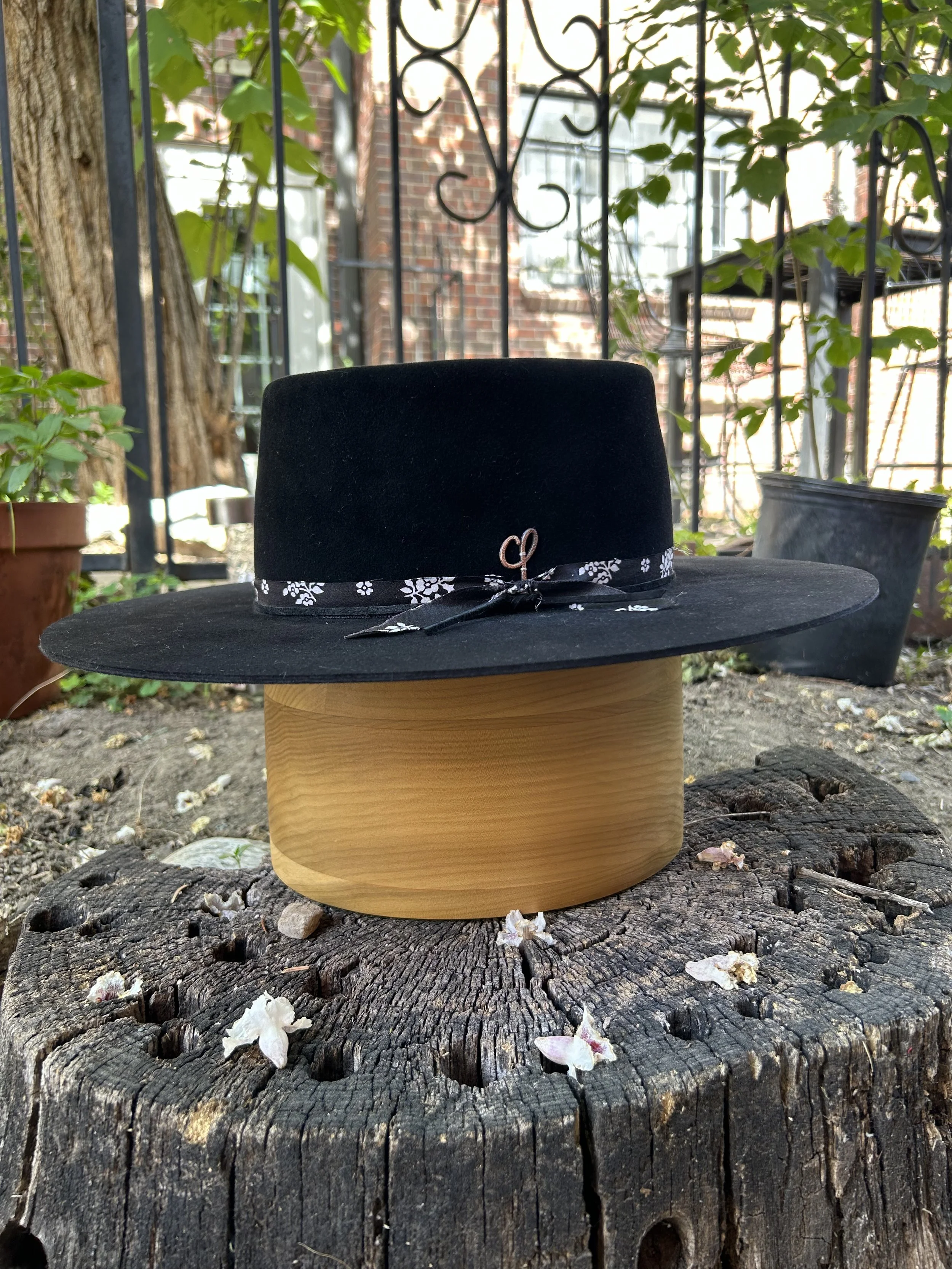 A black wide-brimmed hat with a decorative ribbon and a small metal heart charm, placed on a wooden stump outdoors, with potted plants and a brick building in the background.
