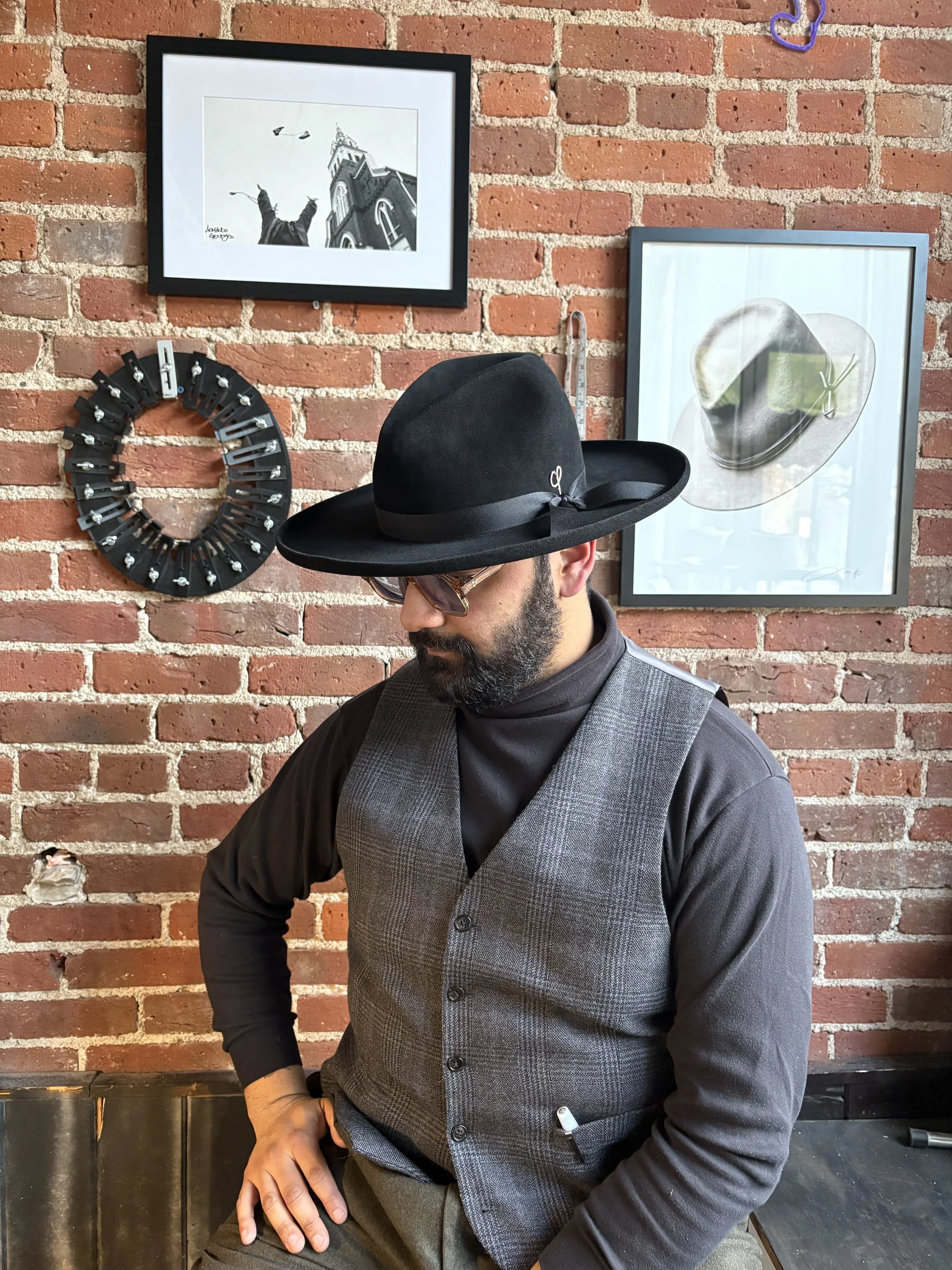 Man wearing a wide-brimmed black hat, glasses, a black turtleneck, and a gray vest sitting against a brick wall with framed photographs and artwork behind him.