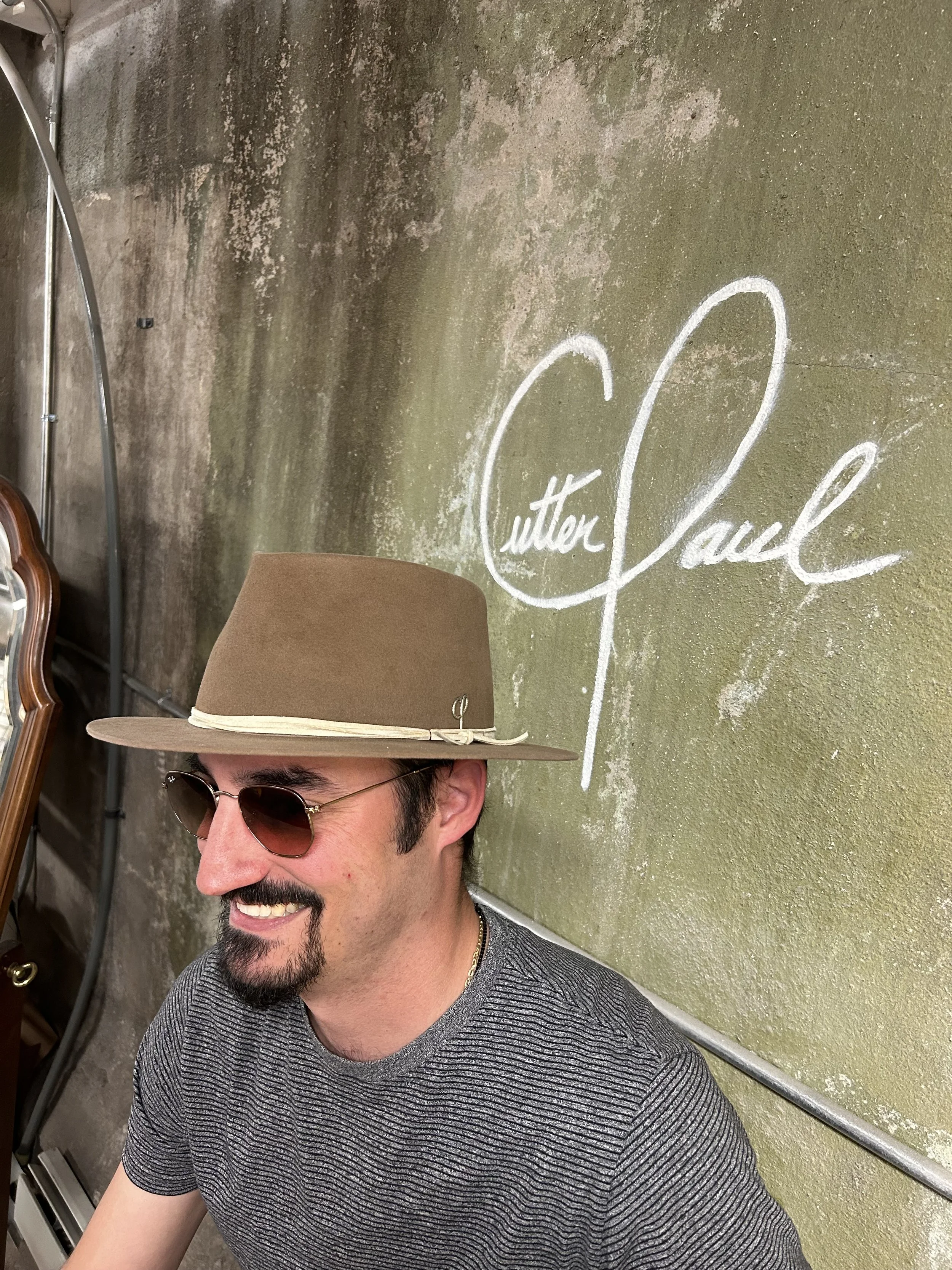 A smiling man wearing sunglasses, a brown hat, and a striped shirt, sitting in front of a textured green wall with the words 'Cutter Lounge' written in white chalk.