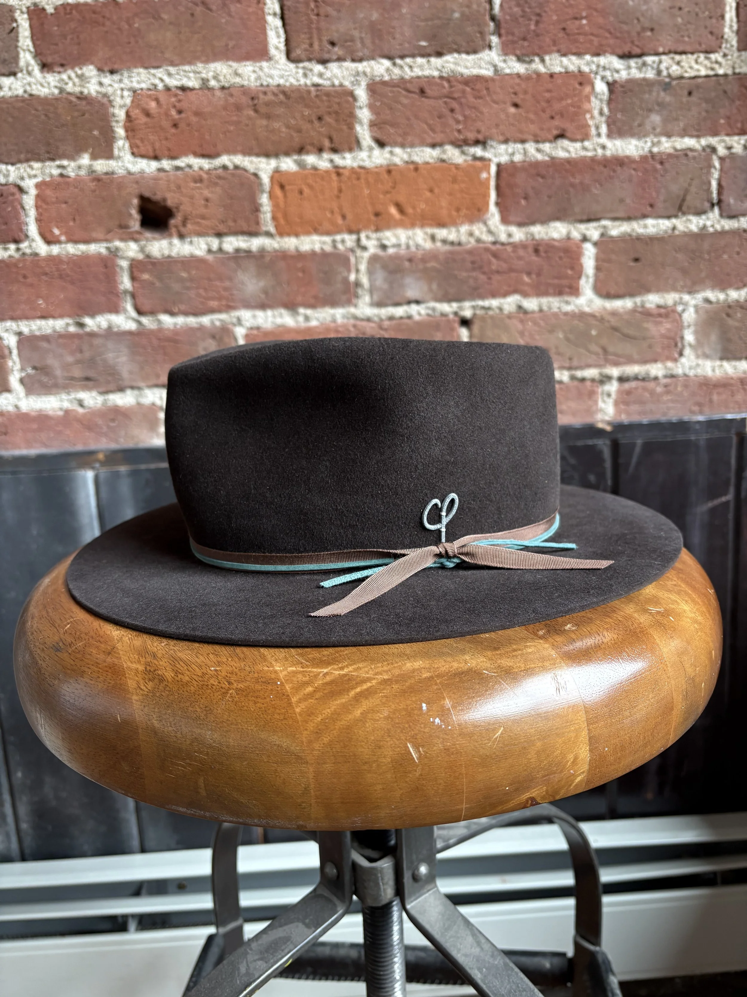 A black felt hat with a brown ribbon and teal accents, resting on a round wooden table in front of a brick wall.
