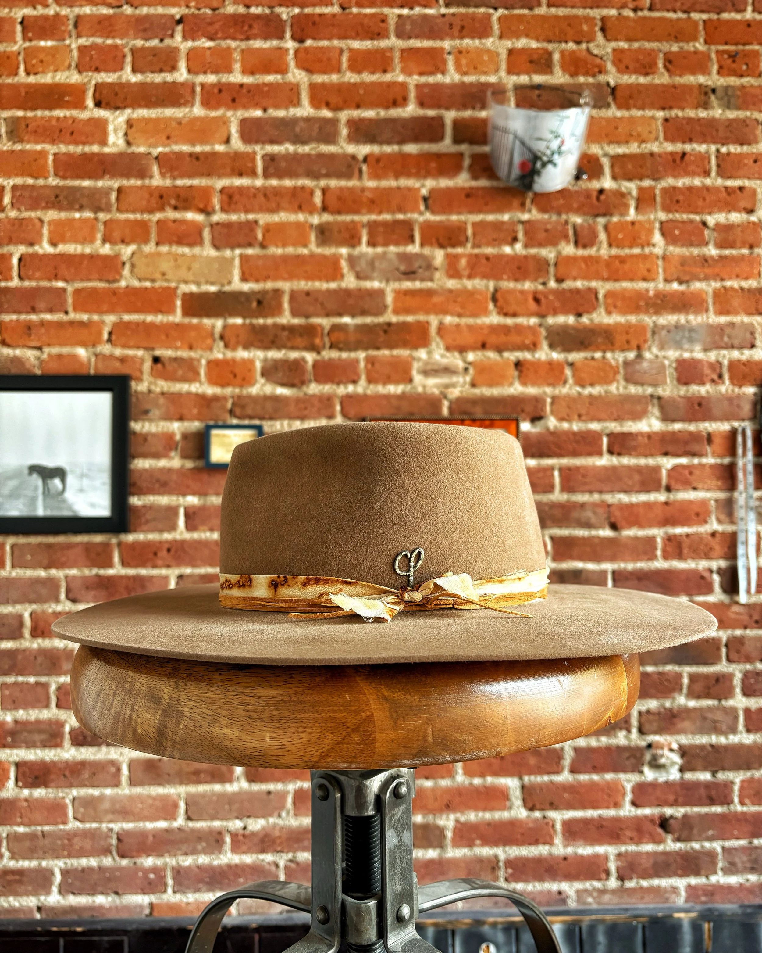 A tan wide-brimmed hat with a ribbon, placed on a round wooden table in front of a red brick wall.
