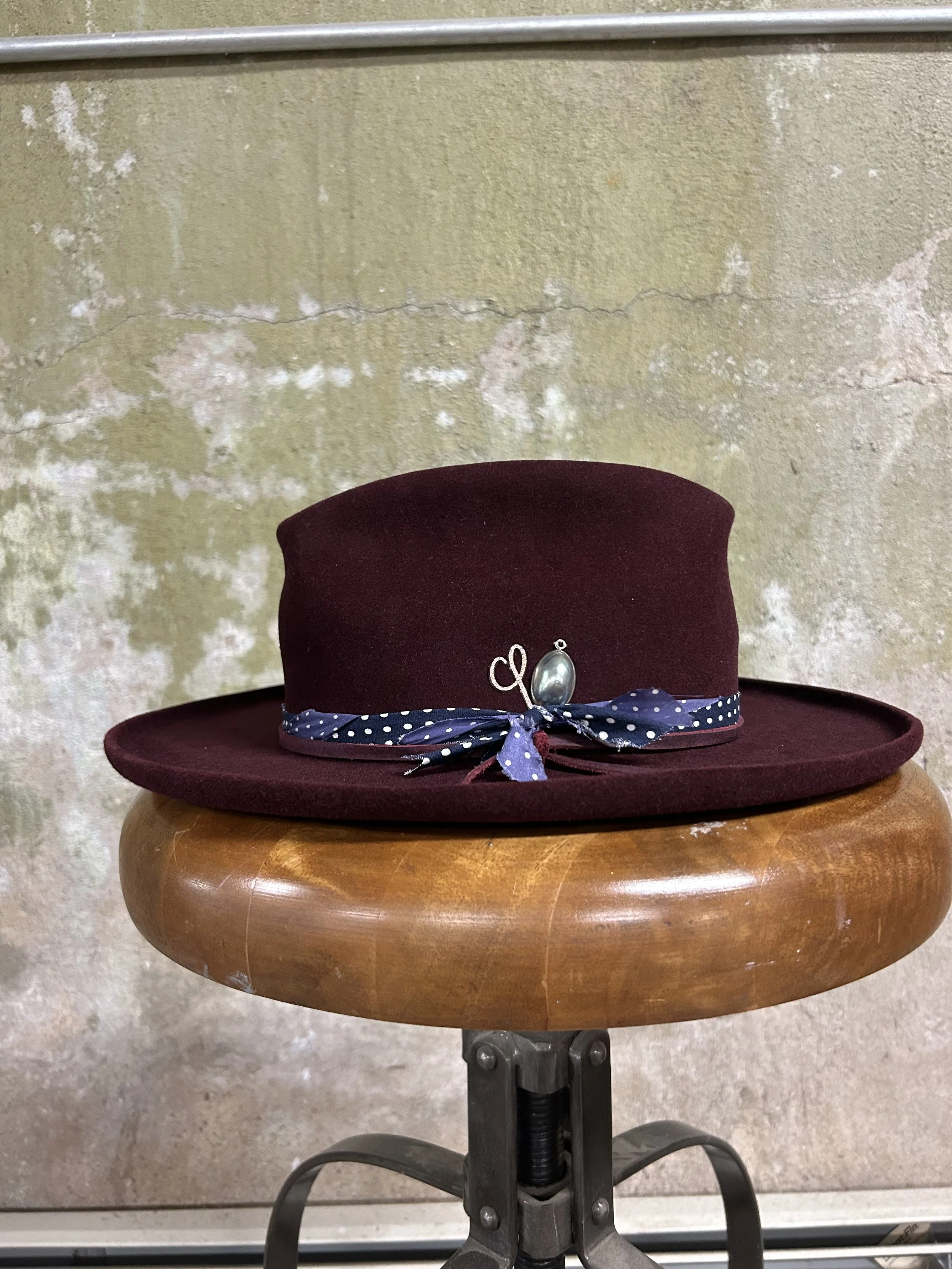 A burgundy felt hat with a blue polka dot ribbon and a decorative pin, placed on a wooden stool against a textured, weathered wall background.