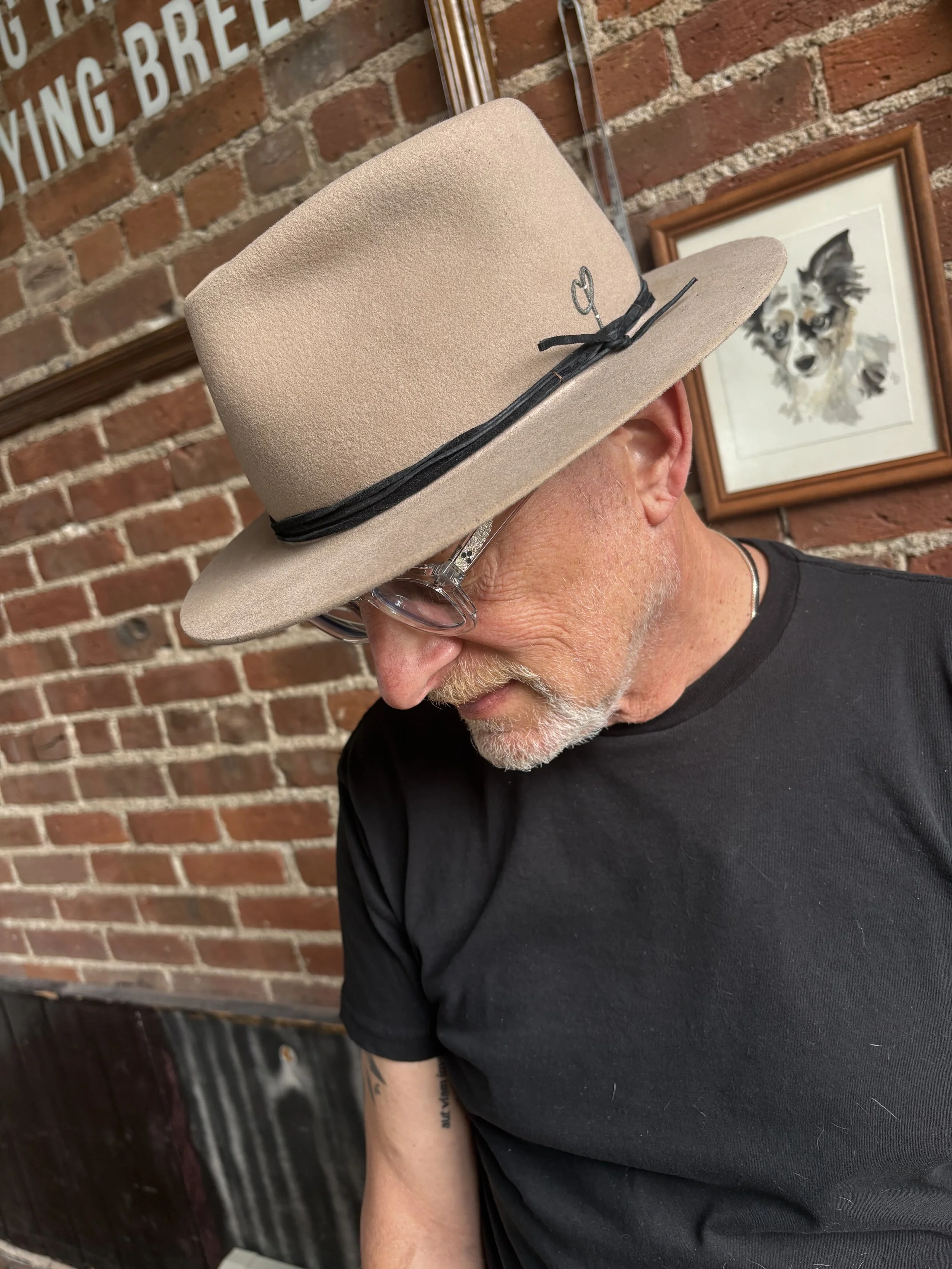 A man with white hair and a beard wearing glasses and a beige fedora hat decorated with a black band and small black bow, looking down, in front of a brick wall with framed artwork of a dog.