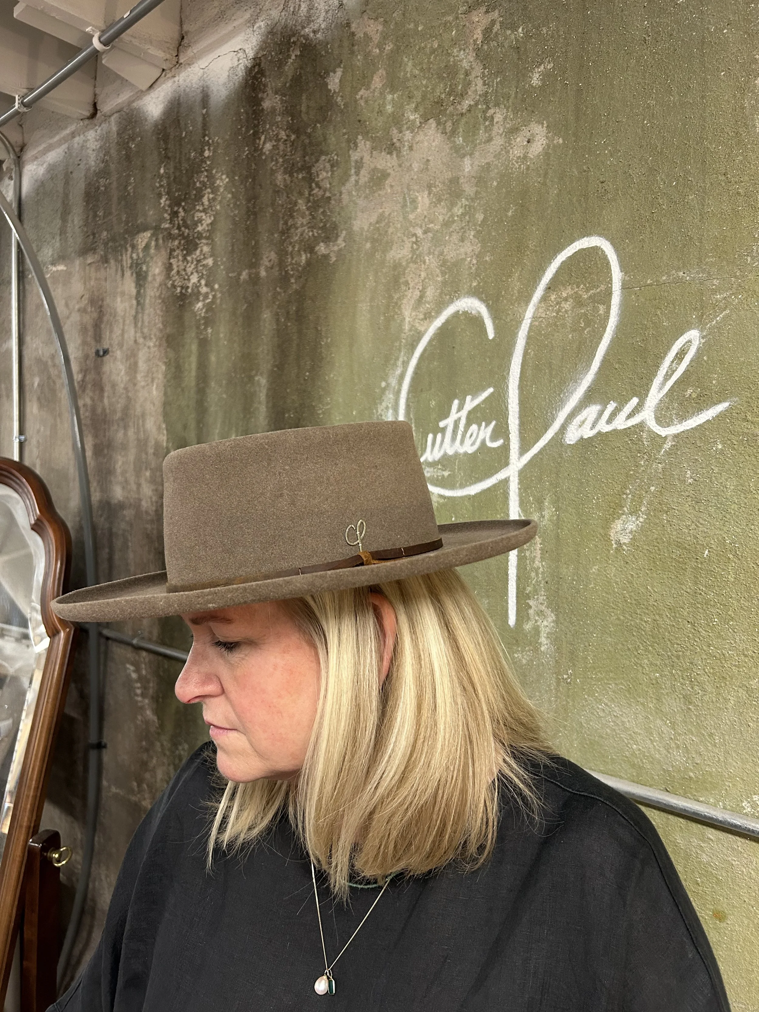 A woman with blonde hair wearing a brown felt hat with a small decorative bow, standing in front of a textured moss-covered wall with white cursive writing that reads 'Sutton Paul'.