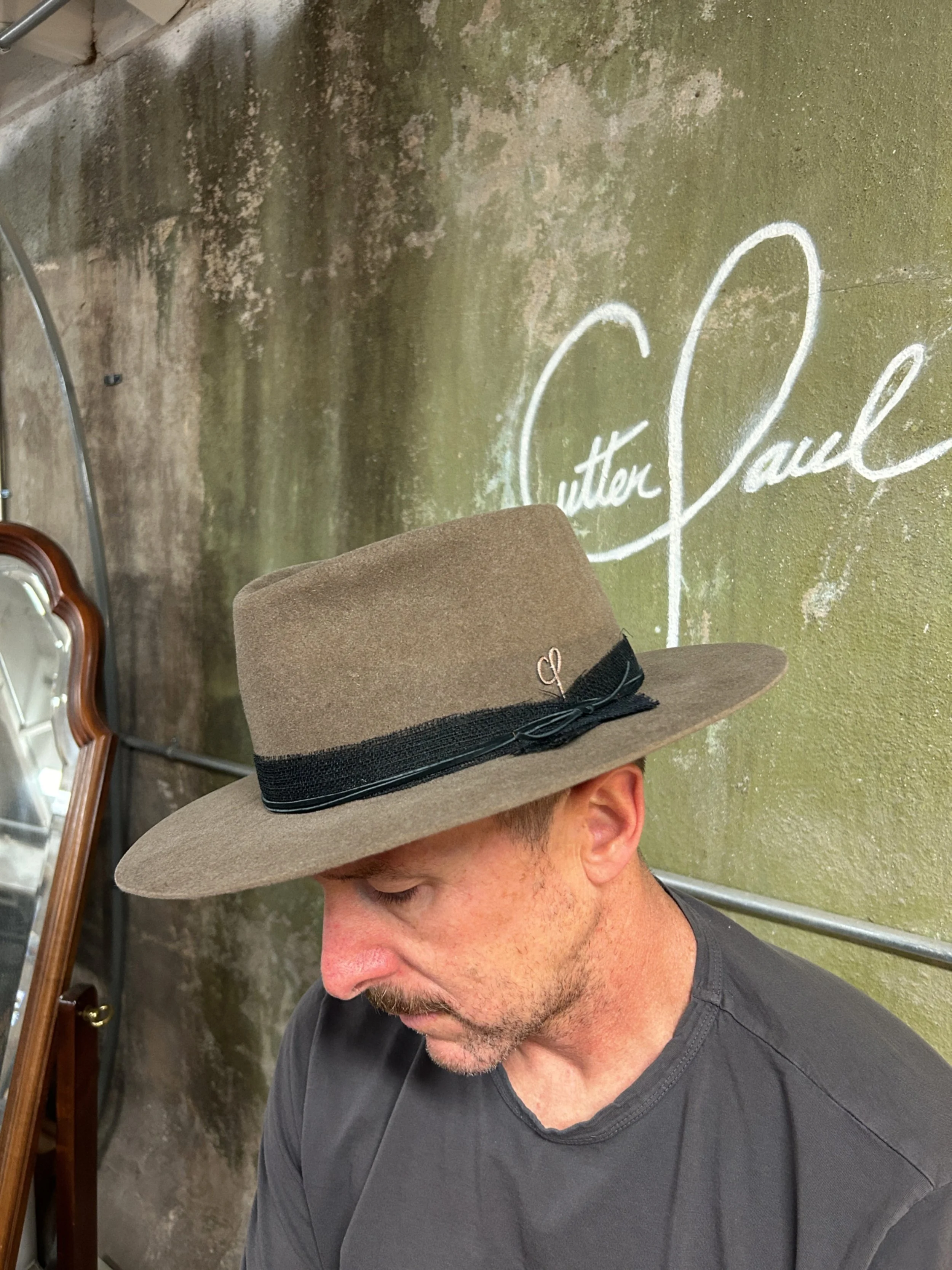 A man wearing a wide-brimmed brown hat with a black band, sitting with his head bowed in front of a distressed green wall with white signature-style writing that reads "Cutter & Co.".