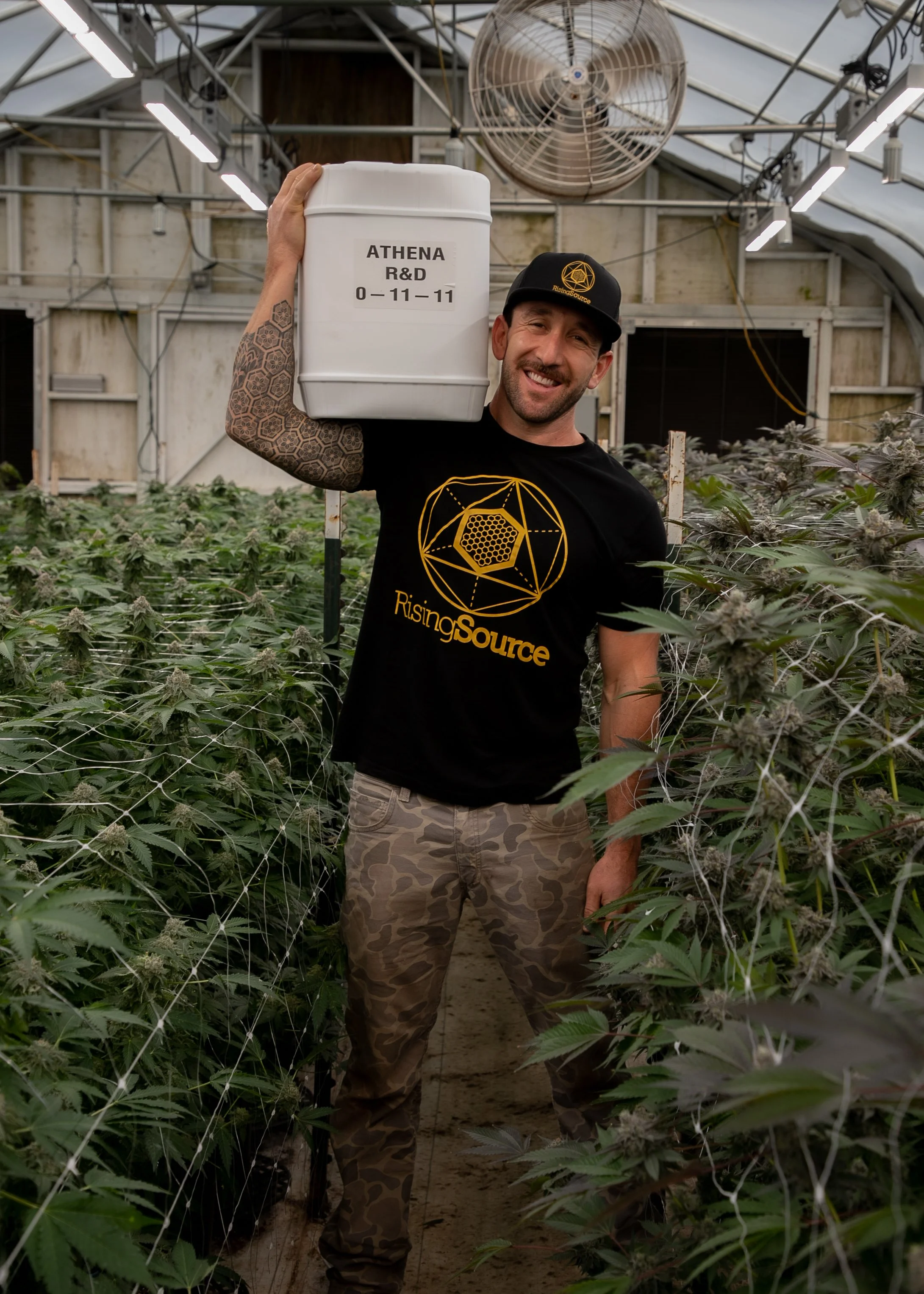 A smiling man in a black RisingSource T-shirt and camouflage pants standing in a greenhouse filled with cannabis plants, holding a white container labeled Athena R&D.