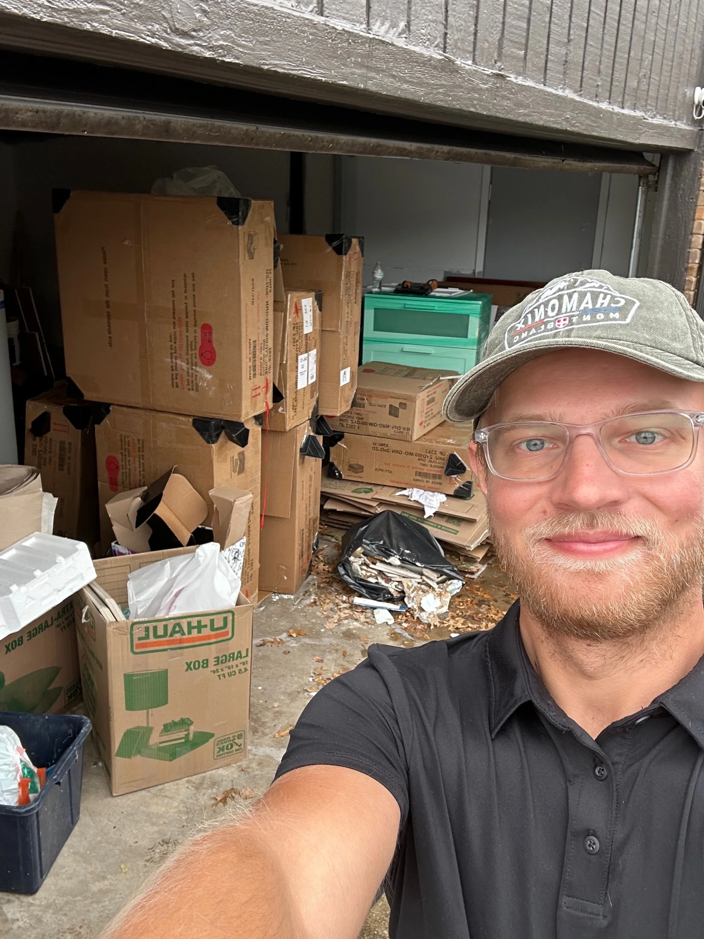 A man taking a selfie in front of a garage filled with cardboard boxes, paper, and trash, with a green storage container and tools visible in the background.