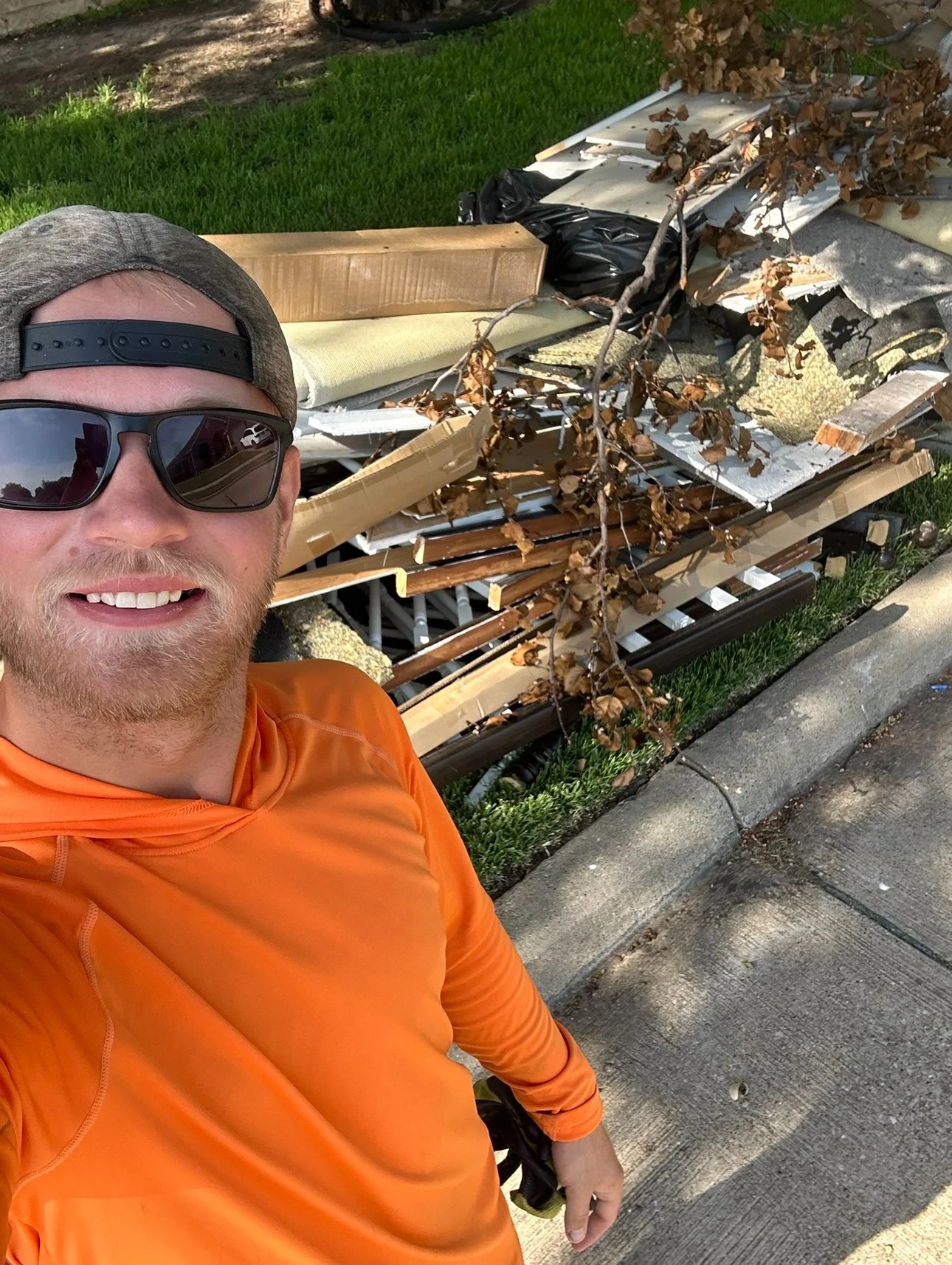 A man wearing sunglasses and an orange athletic shirt taking a selfie next to a pile of debris on the sidewalk, including wood, metal, insulation, a branch with brown leaves, and other construction materials.