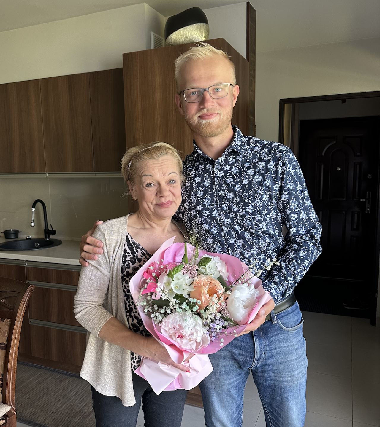 A young man and an older woman standing together indoors, holding a colorful bouquet of flowers, both smiling. The woman is wearing a beige cardigan and the man is wearing glasses and a floral shirt.