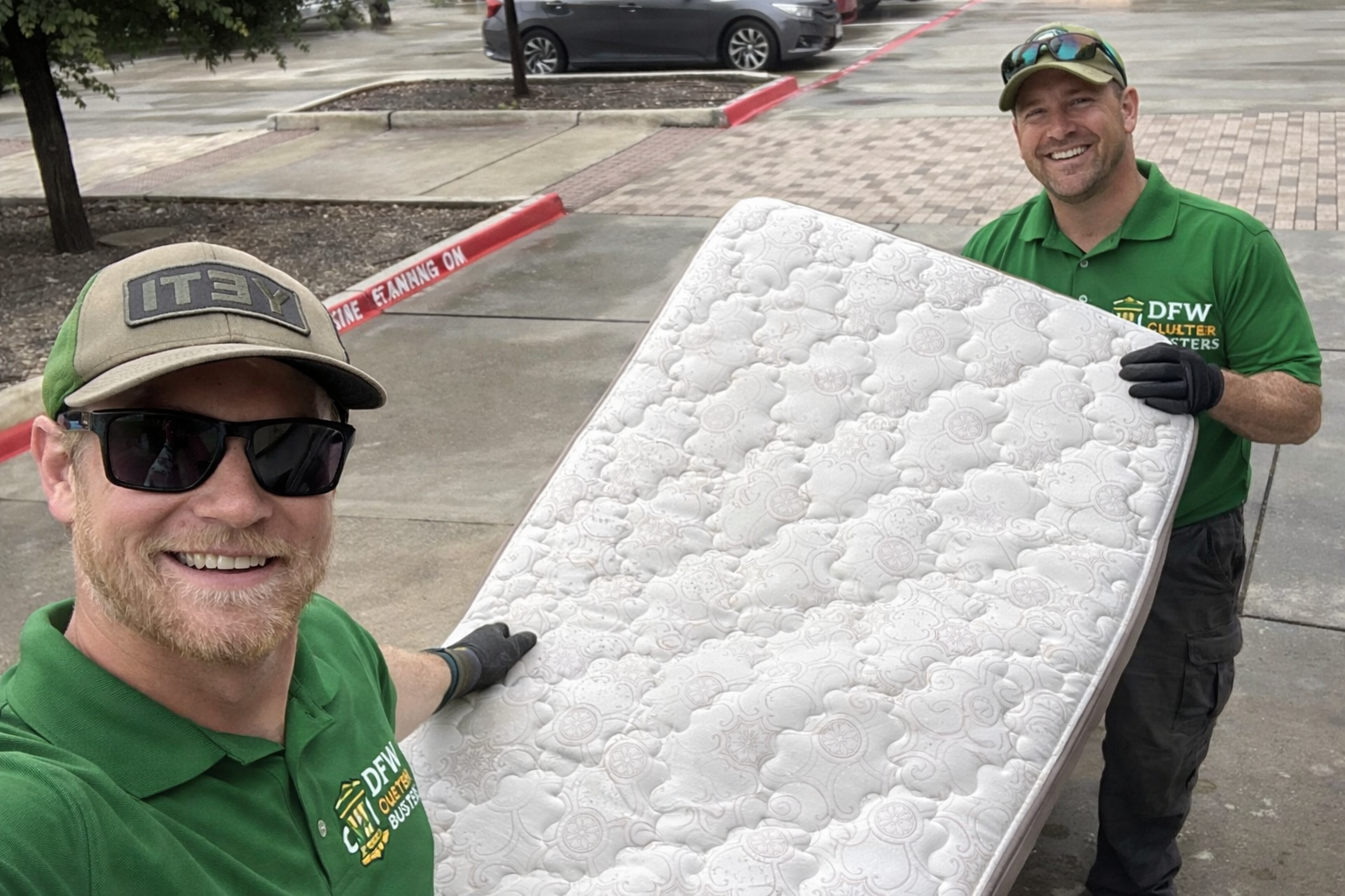 Two men wearing green shirts and gloves are carrying a mattress outside of a building. One man is taking a selfie, and both are smiling.
