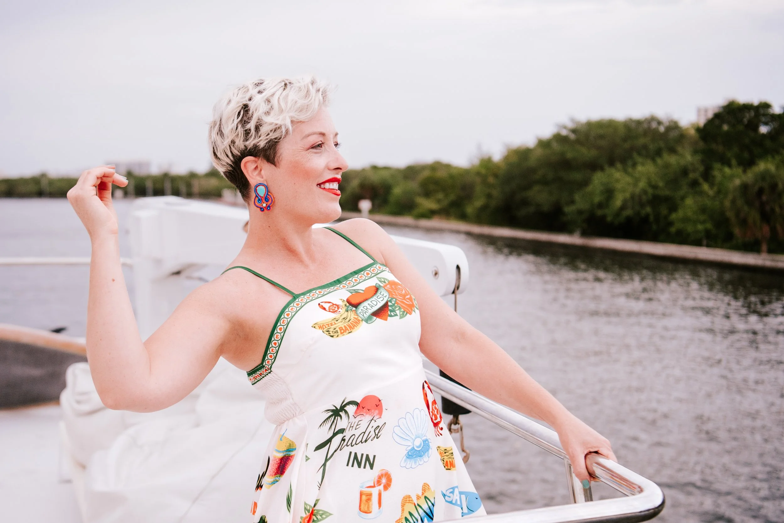 A woman with short, curly, gray hair, wearing red lipstick, and colorful earrings, is standing on a boat by a river, smiling and enjoying the scenery, with trees and a cloudy sky in the background.