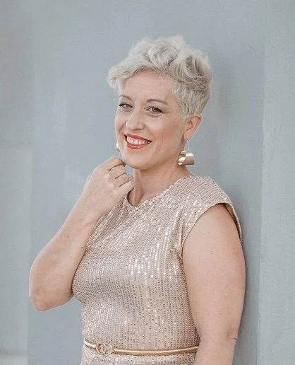 A smiling woman with short, curly white hair, wearing a sleeveless, shimmering beige dress with a gold belt, standing against a light blue background.