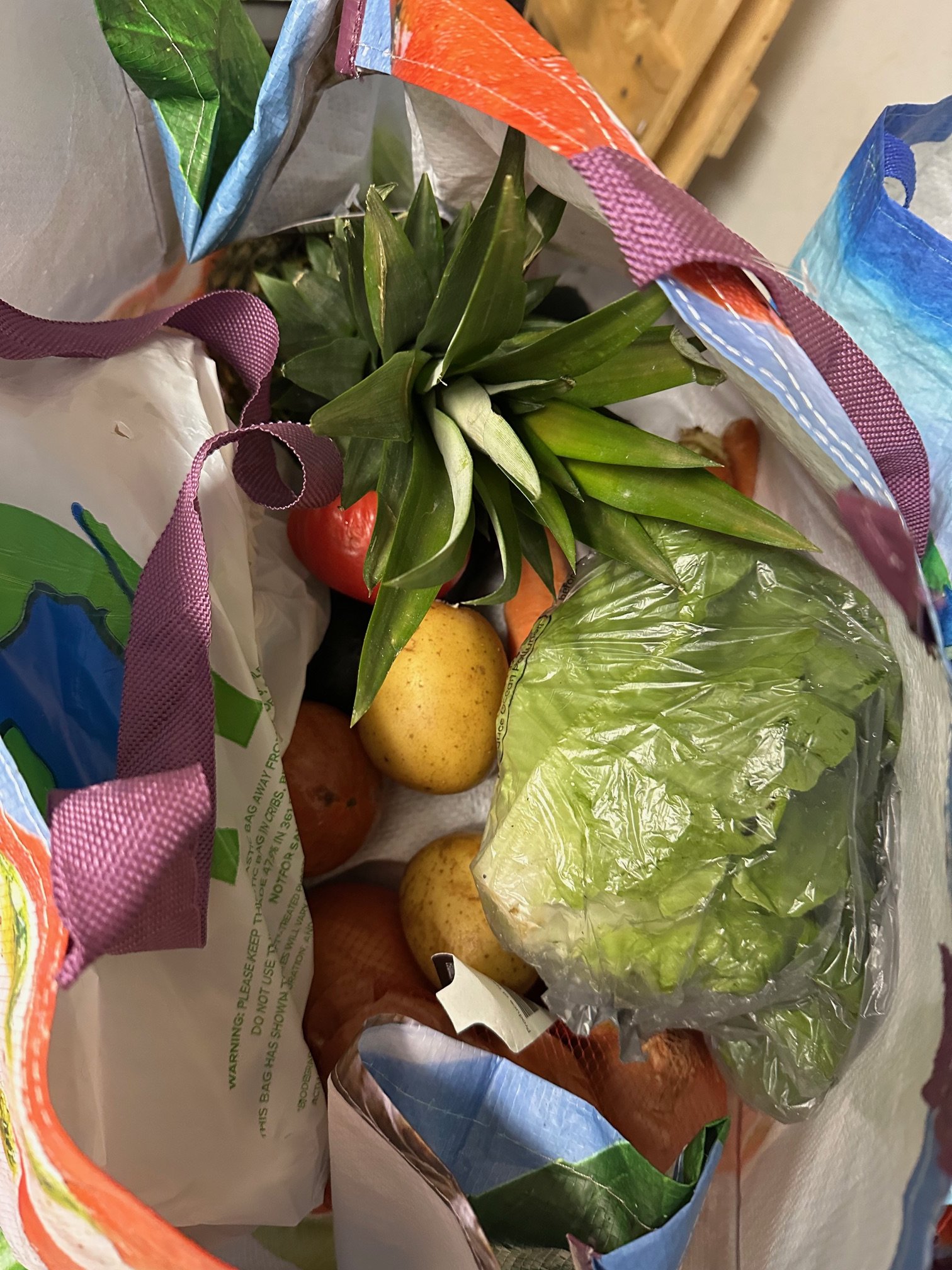 Top view of a reusable shopping bag filled with various fresh fruits and vegetables, including a pineapple, apples, lettuce, carrots, and a pear.