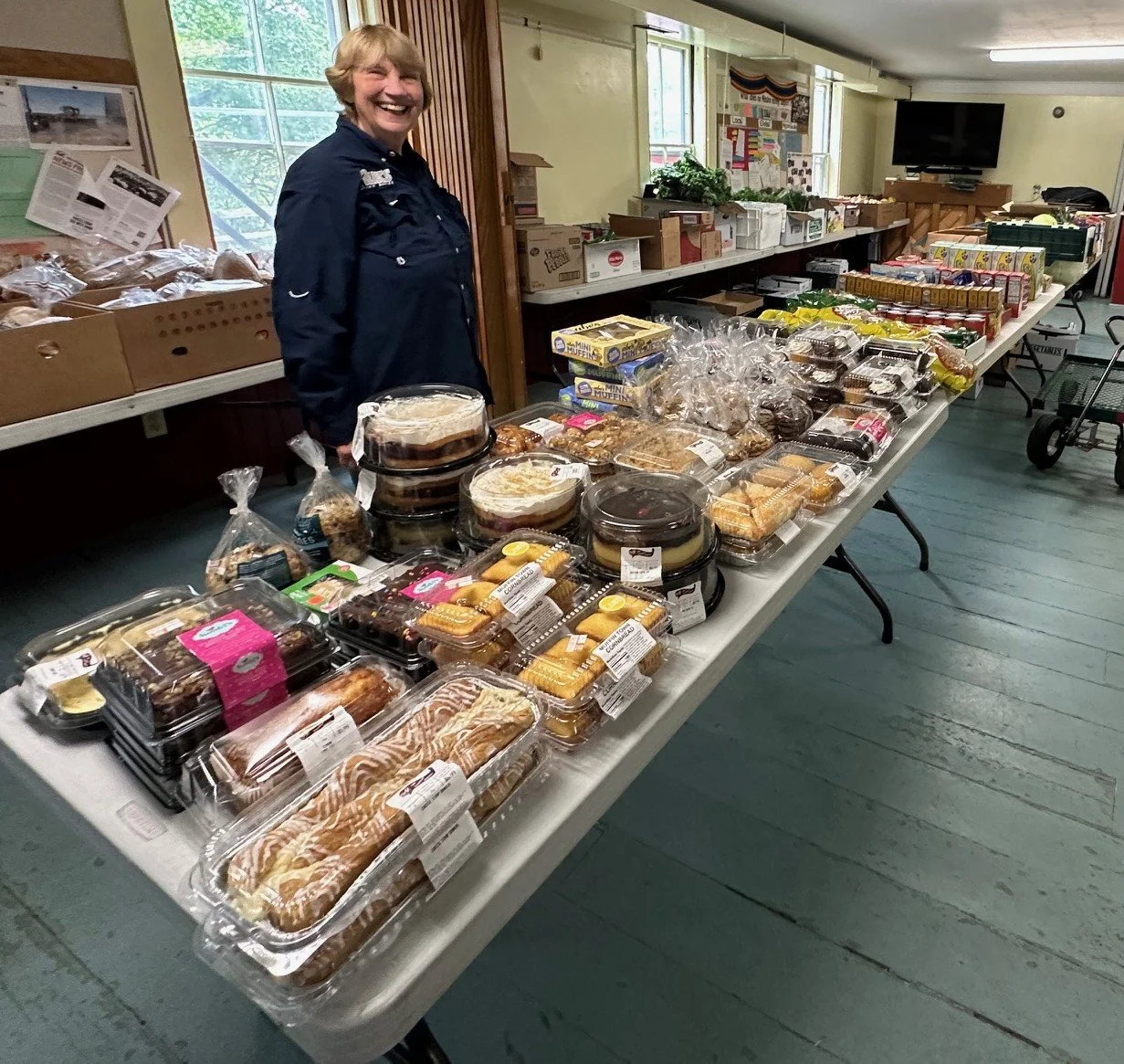 A smiling woman stands behind a long table filled with baked goods, including cakes, cookies, and pastries, in an indoor setting with windows and bulletin boards in the background.