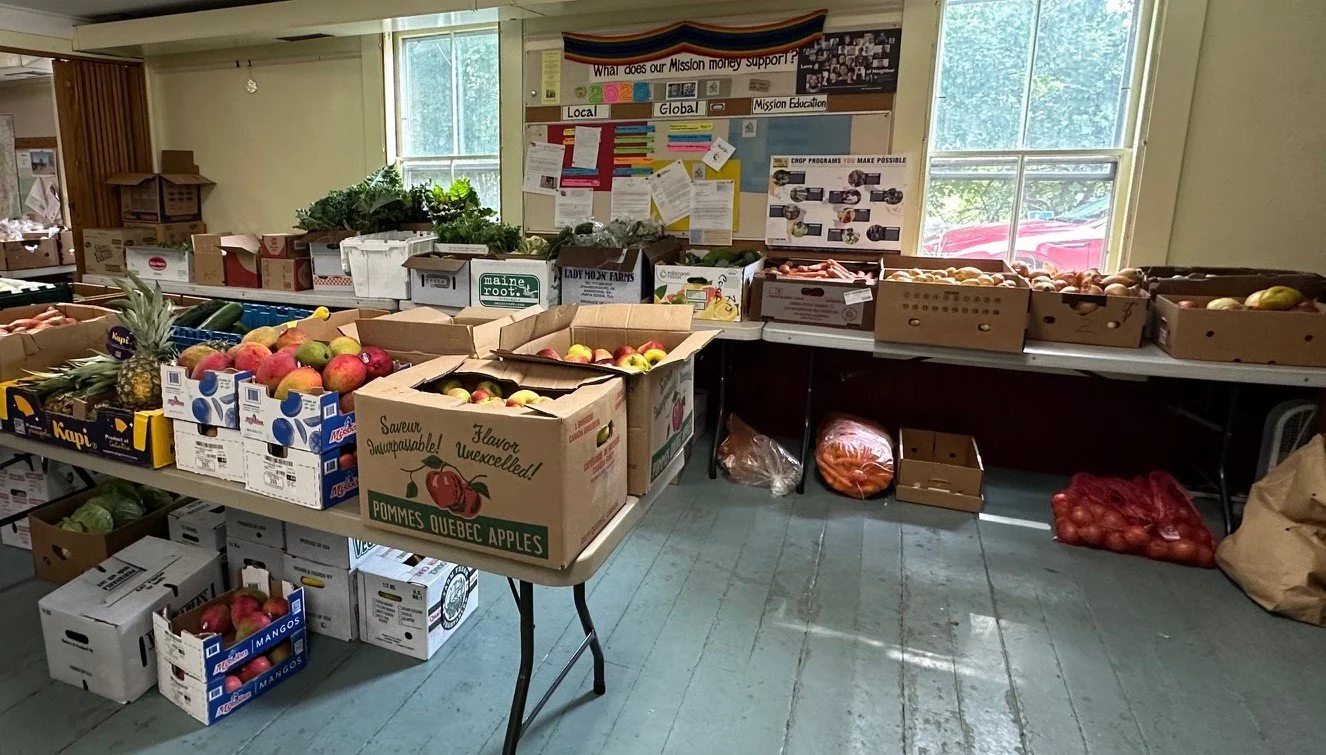 Indoor produce stand with boxes of apples, onions, pineapples, and greens, set against a wall with informational posters and large windows allowing natural light.