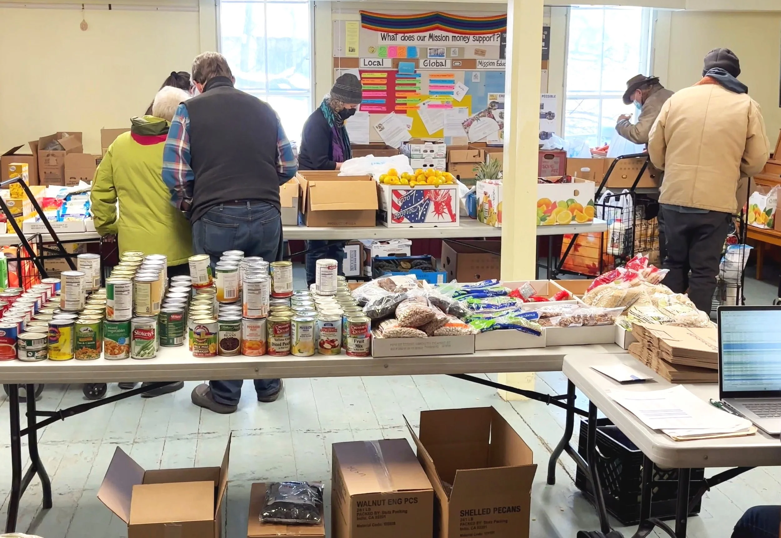 People volunteering at a food bank, sorting and packing canned goods, fresh produce, and other food items on tables in a room with large windows.