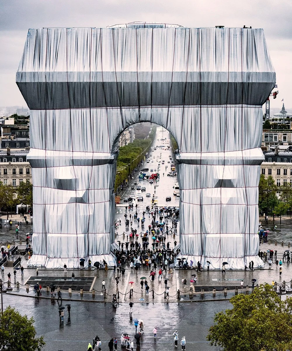 A large building under construction covered with a gray and white protective fabric, shaped like the Eiffel Tower, with a crowd of people and umbrellas in the plaza below during rainy weather.