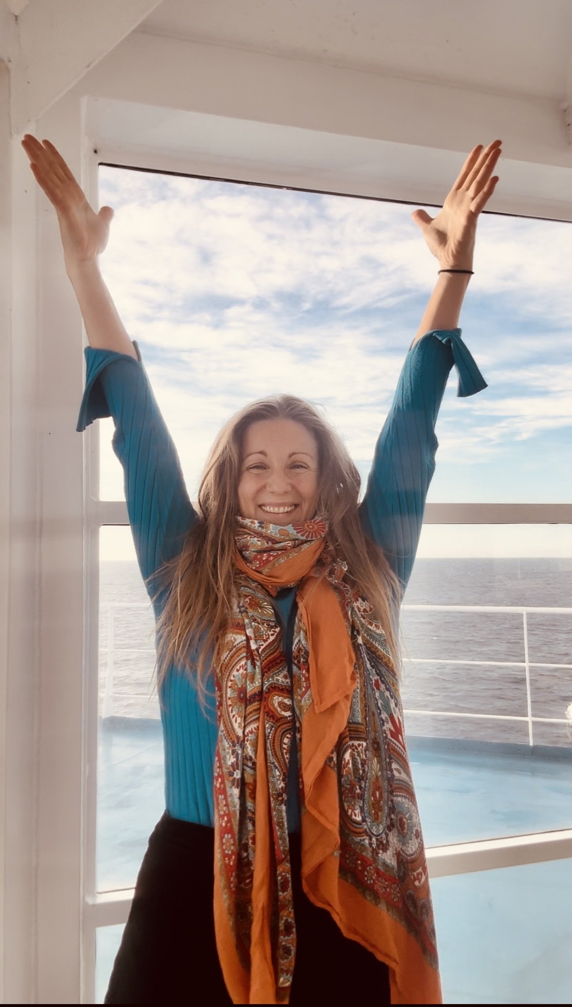 Kvinna med långt hår, som ler brett och sträcker armarna uppåt, står framför ett stort fönster med havet i bakgrunden, bär en blå tröja och en orange scarf. Petra Koppla Dahlberg